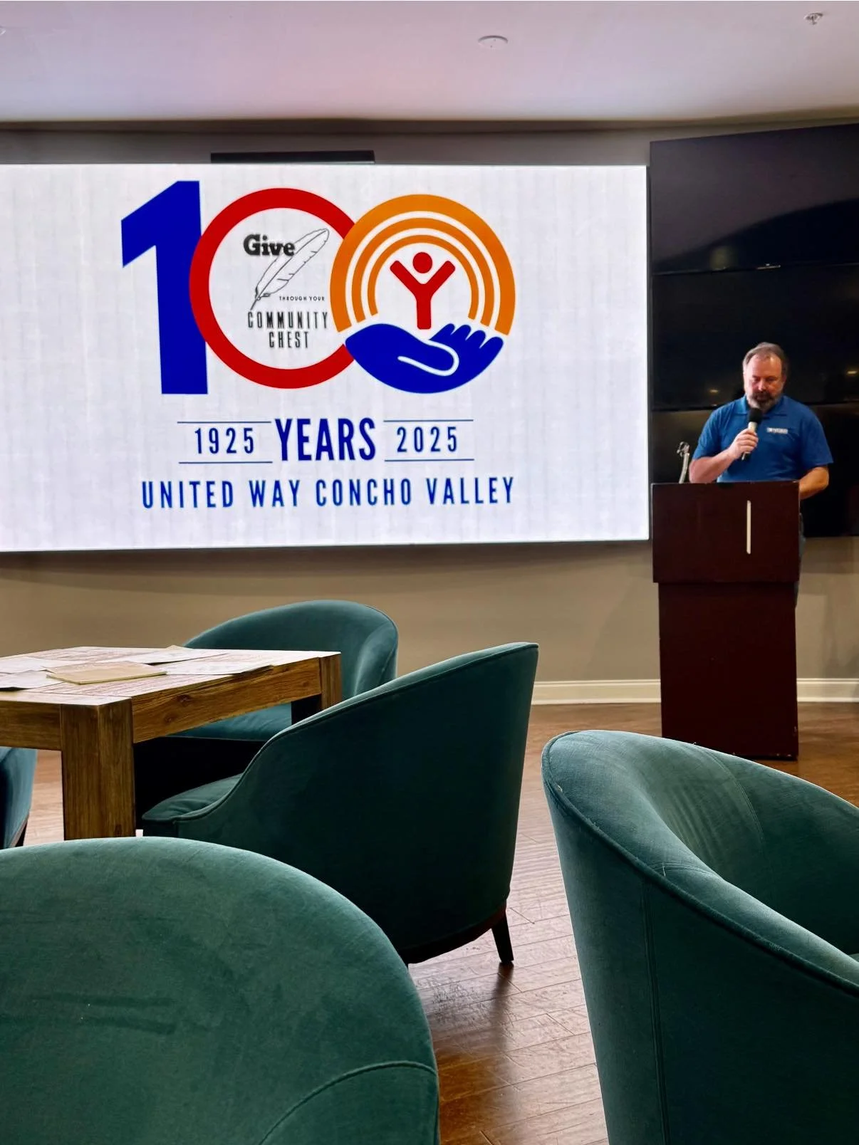 Presentation screen displaying the logo of United Way Concho Valley celebrating 100 years from 1925 to 2025 with a man speaking at a podium and seating area in front.