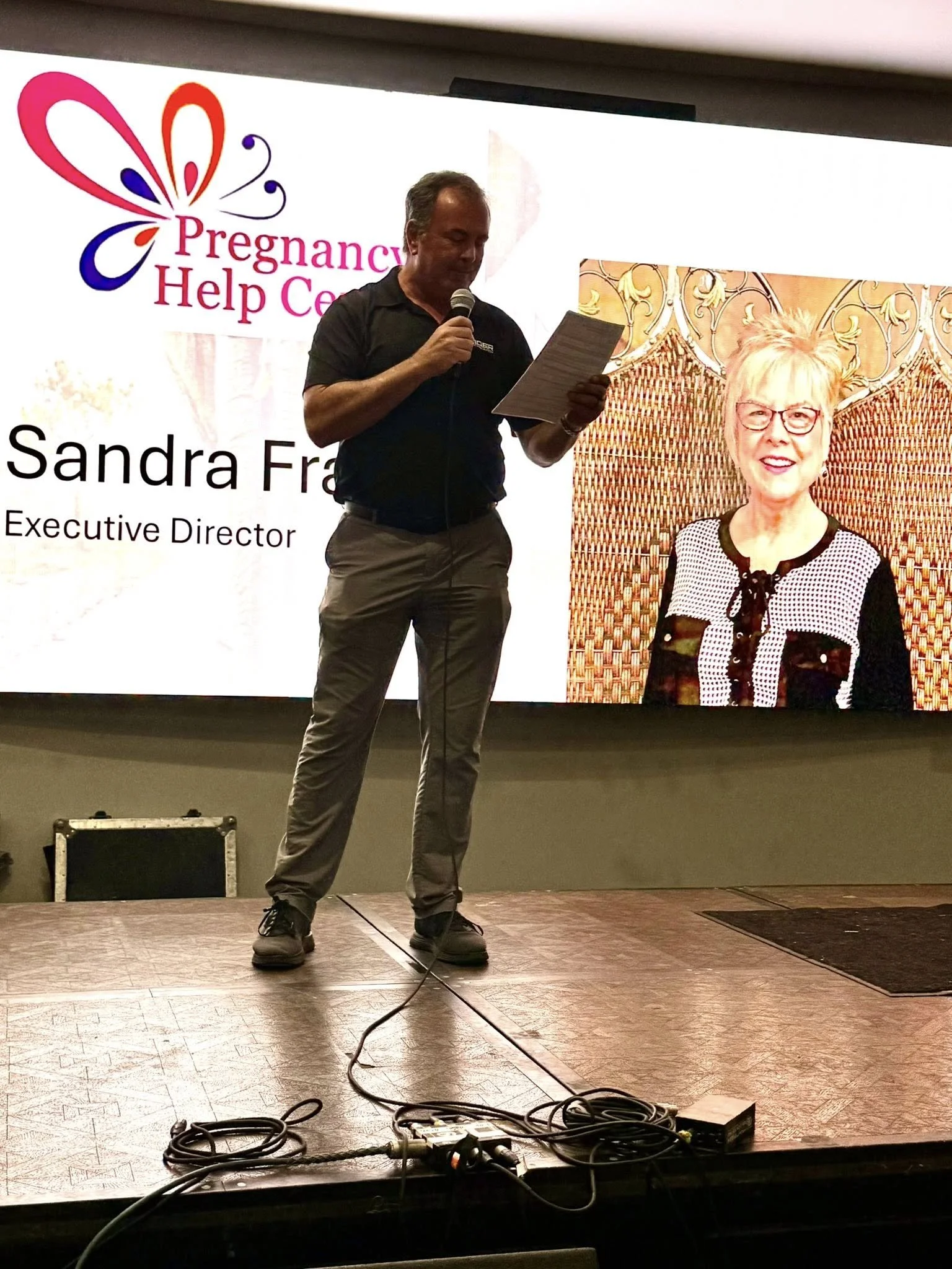 A man standing on a stage reading from a paper while speaking into a microphone, with a large screen behind him displaying a logo for Pregnancy Help Center, a photo of a woman, and the text 'Sandra F' and 'Executive Director'.