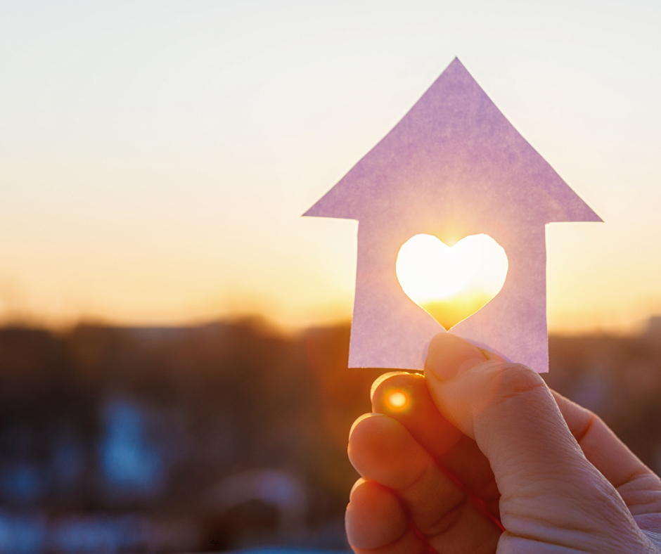 Hand holding a paper house cutout with a heart-shaped opening at sunset