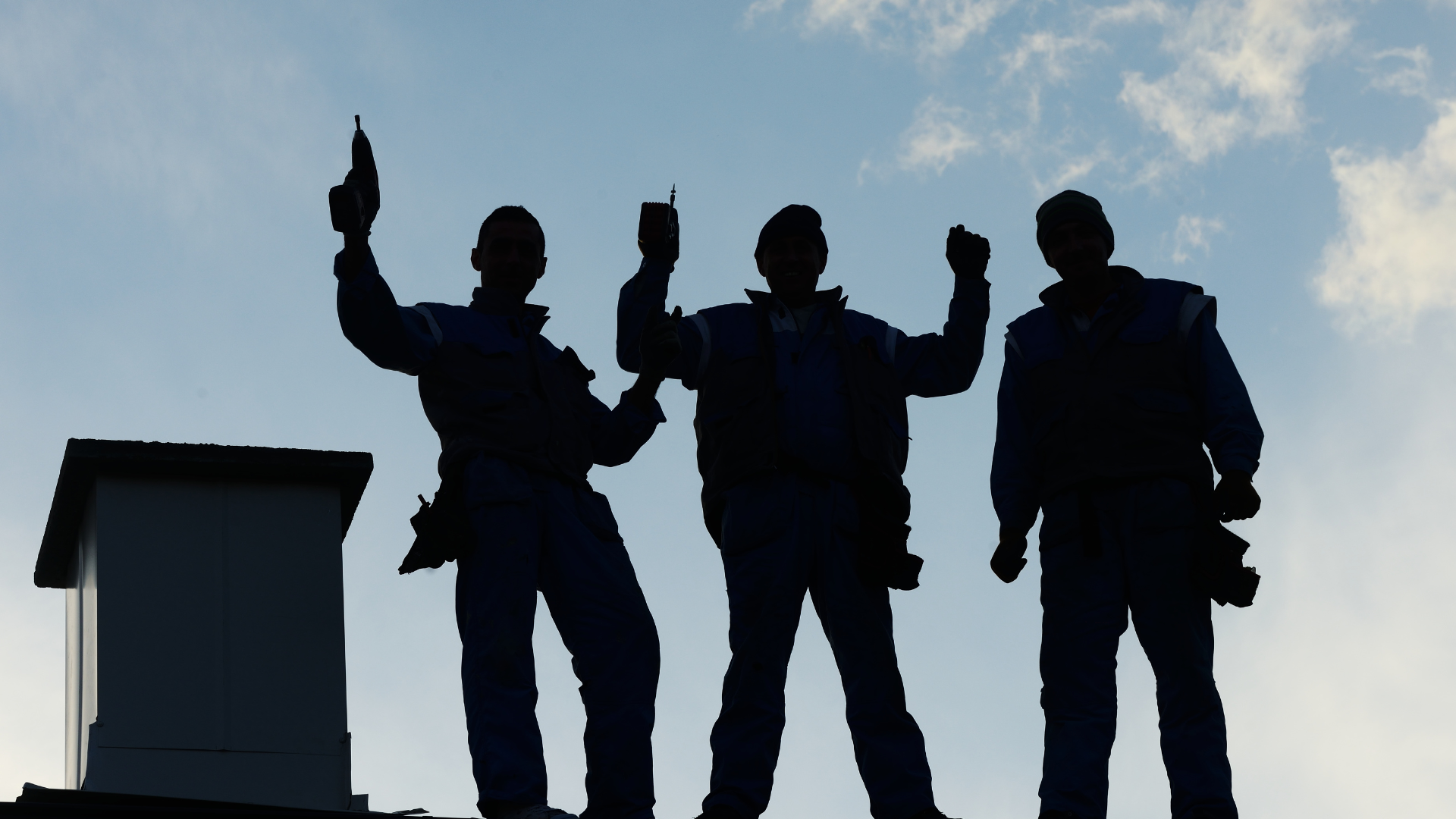 Silhouettes of three construction workers standing on a roof with a skylight and clear sky in the background.