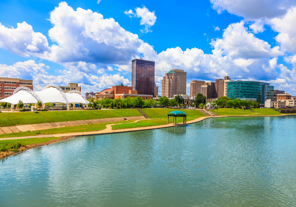 Dayton City skyline with tall buildings and a park with a pond, green grass, and a pavilion under a blue sky with fluffy clouds.