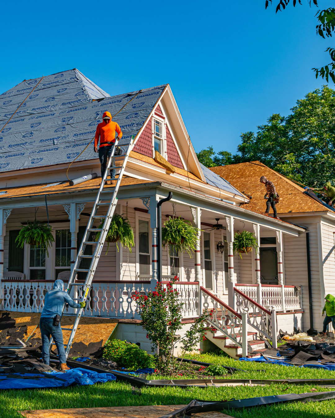 Construction workers replacing the roof of a house, with some working on the shingles and others on the roof, and blue sky in the background.