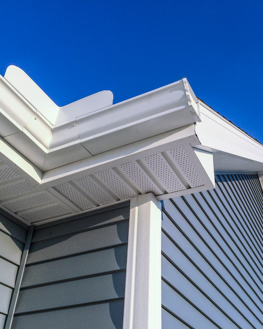 Close-up of the corner of a house with grey siding, white soffit, and a white gutter against a clear blue sky.