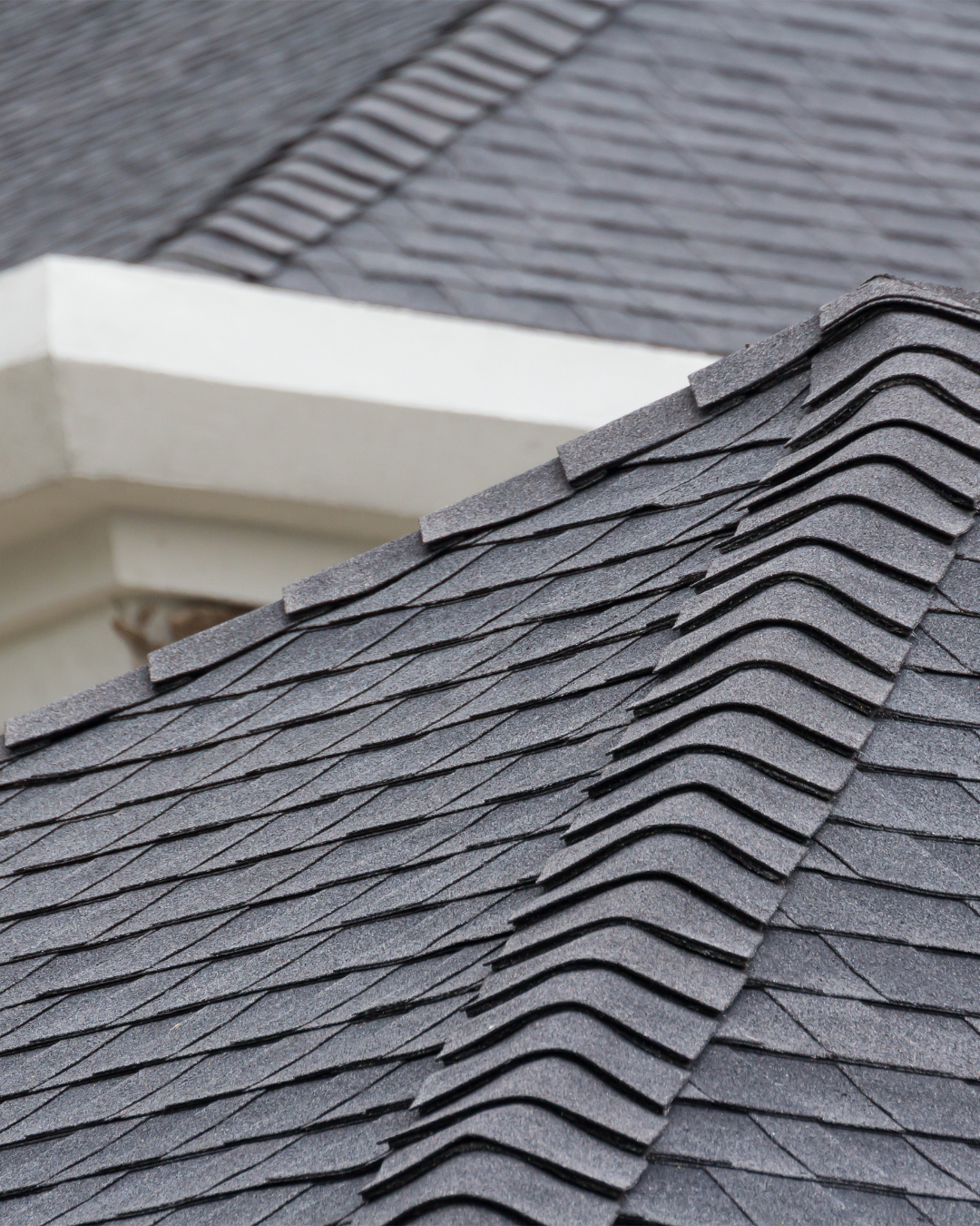 Close-up of dark gray asphalt shingles on a roof with a lighter gray trim and a neighboring roof in the background.