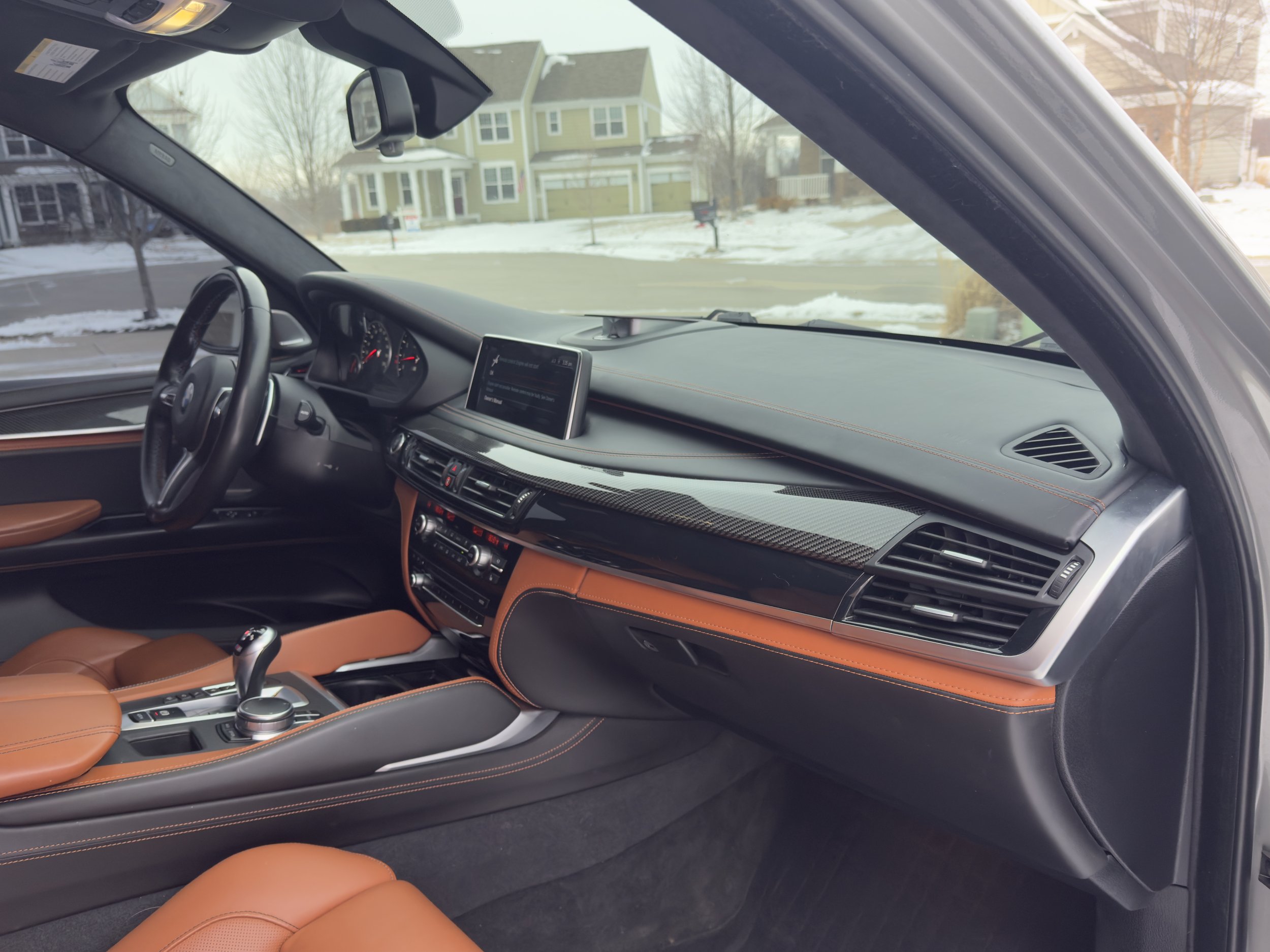 Interior of a luxury car with black and tan leather seats, dashboard, steering wheel, and central console. Outside the window, snow-covered residential neighborhood with houses.
