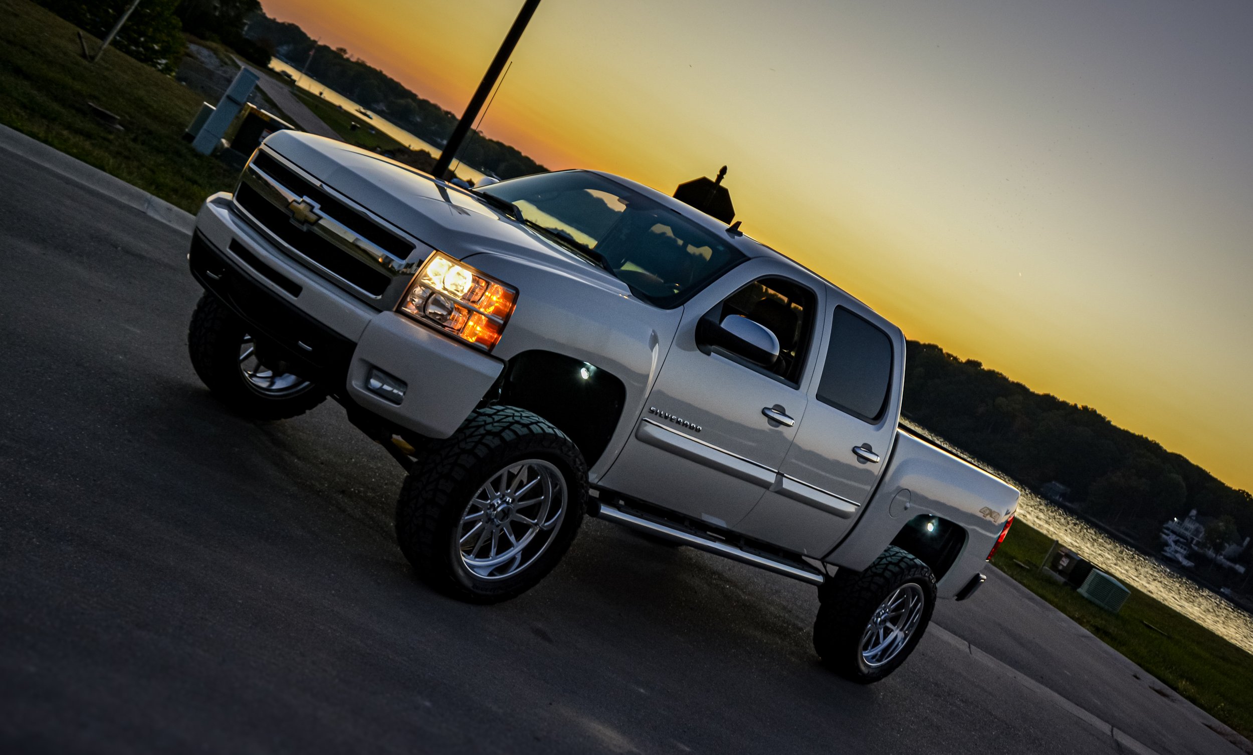 A silver Chevrolet Silverado truck parked outdoors during sunset, with a scenic water view and hills in the background.