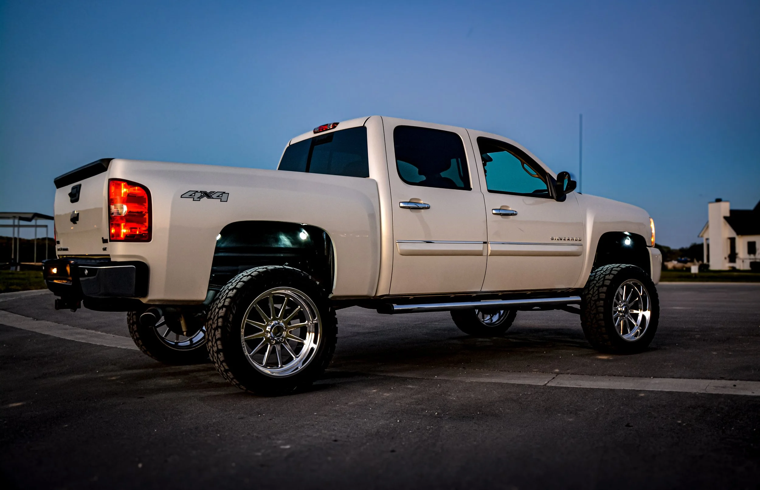A white Chevrolet Silverado pickup truck with four doors, lifted suspension, aftermarket wheels, and tires parked on a paved surface during dusk.