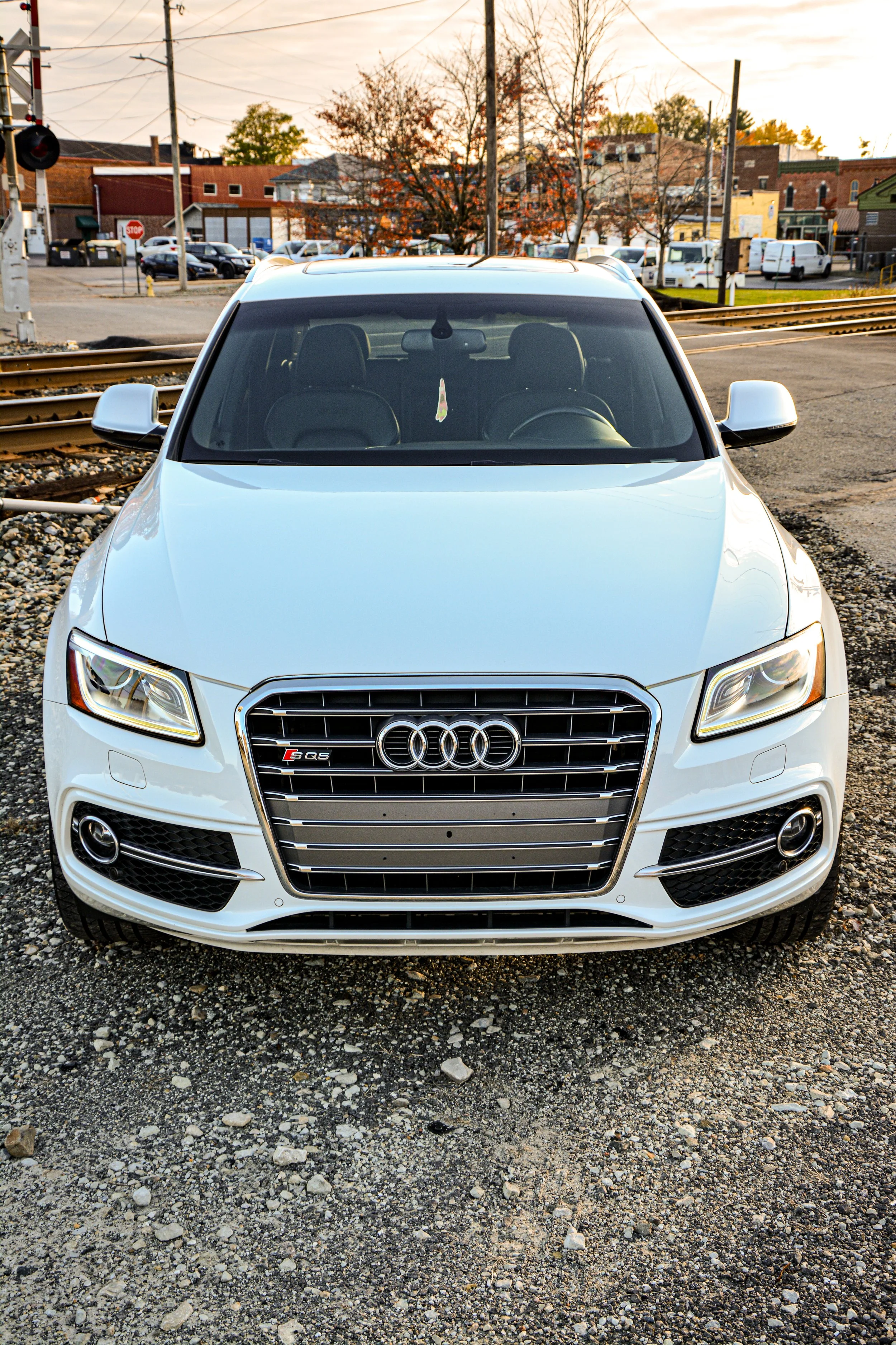 Front view of a white Audi Q5 SUV parked on gravel with railroad tracks behind it, in an urban area during sunset.