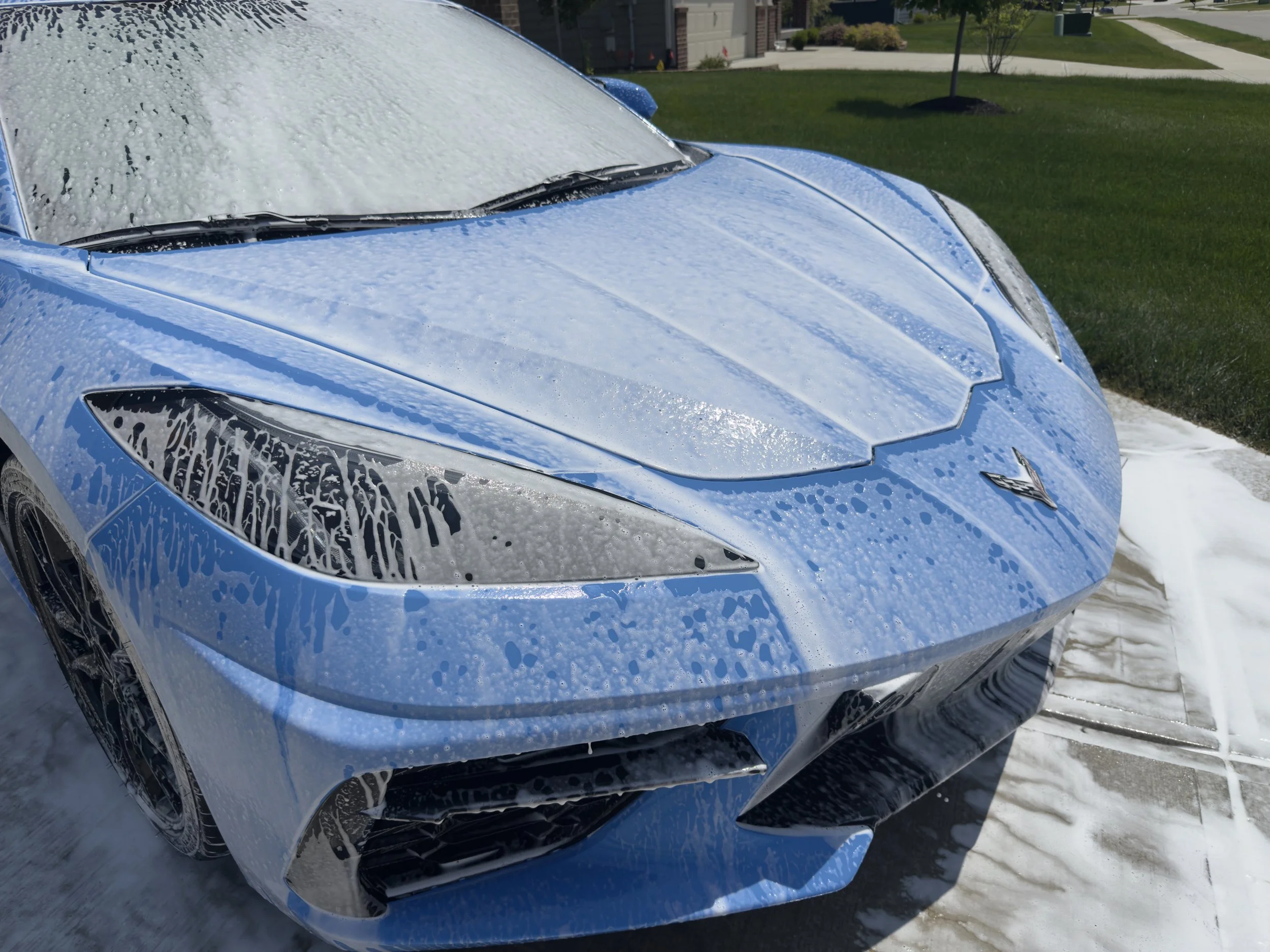 Blue sports car being washed with soap and water in a driveway, with foam on the car's surface and suds on the ground.