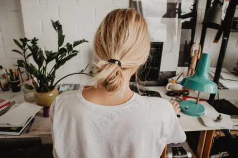 A woman with blonde hair tied back works at a desk in a home office, surrounded by a plant, notebooks, and a blue desk lamp.