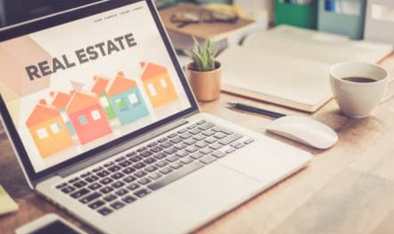 Laptop displaying a real estate website, on a desk with a coffee cup, plant, and office supplies.