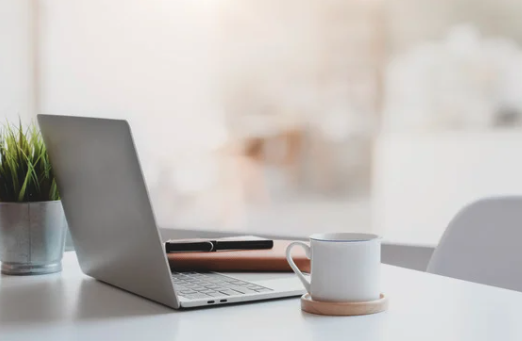 A laptop, a white coffee mug on a wooden coaster, a closed notebook, and a potted plant on a white desk