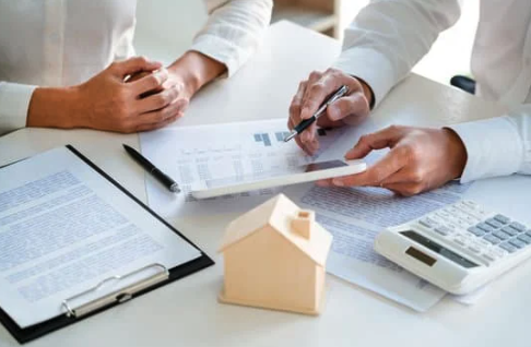 Two people reviewing documents on a desk with a small wooden house model, a calculator, a pen, and a clipboard with papers.