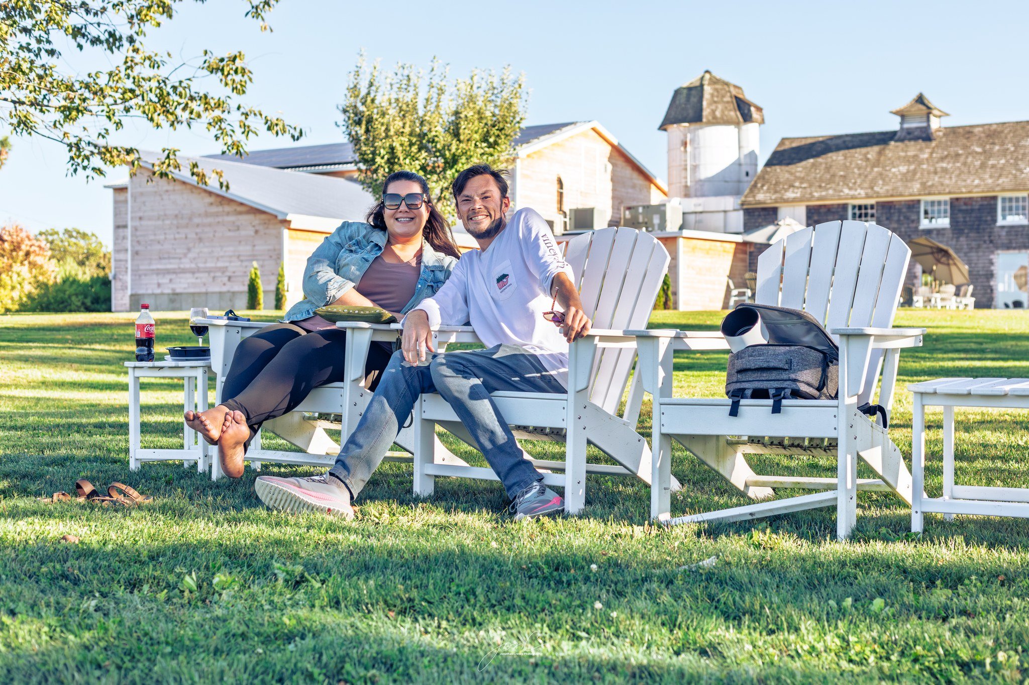 Two friends sitting on white Adirondack chairs outdoors on a grassy lawn, smiling at the camera. One is a woman with dark hair and sunglasses, the other is a man with a beard. There are trees, buildings, and a clear sky in the background, with a bottle of soda and a backpack nearby.