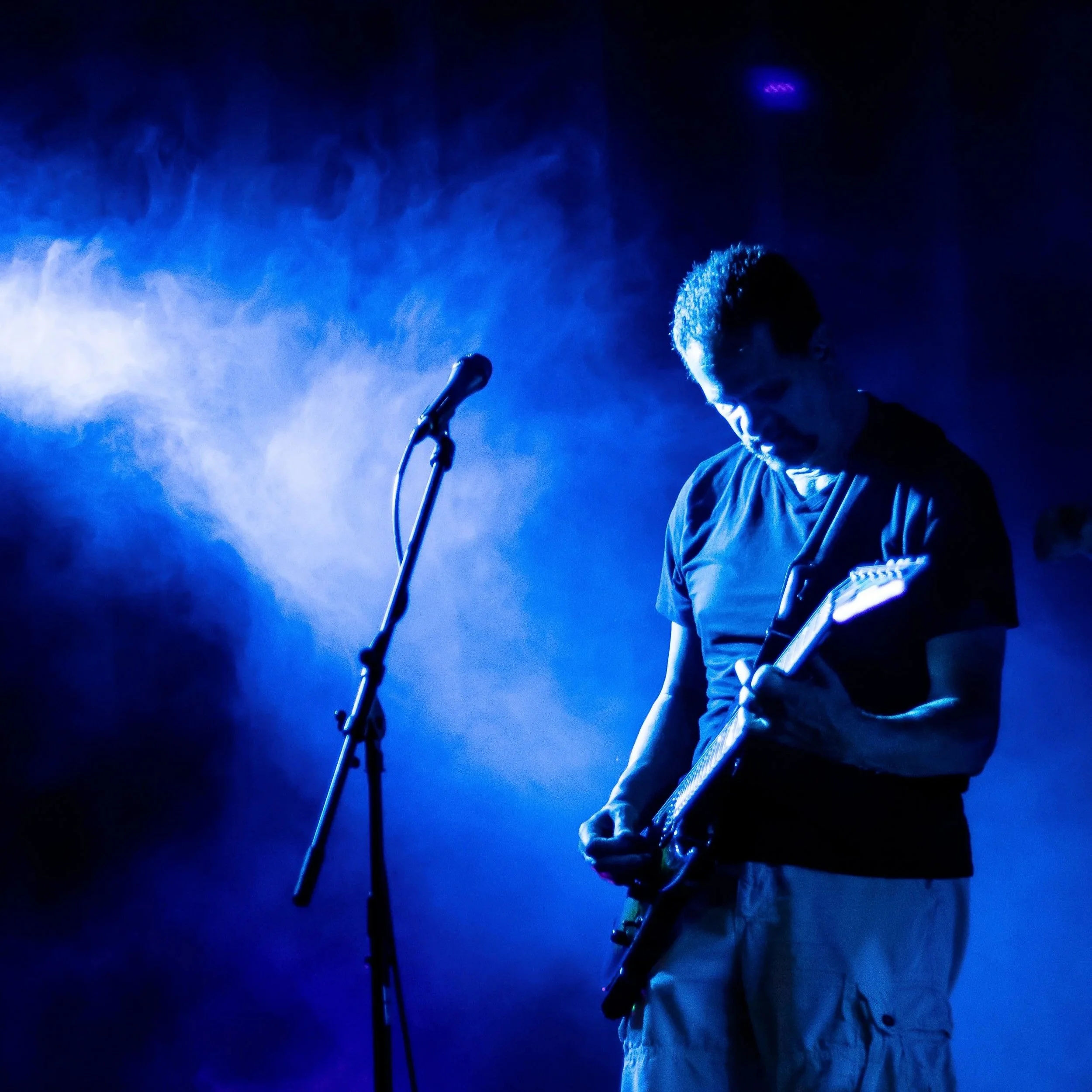 A person playing an electric guitar on stage with blue lighting and smoke, standing near a microphone.