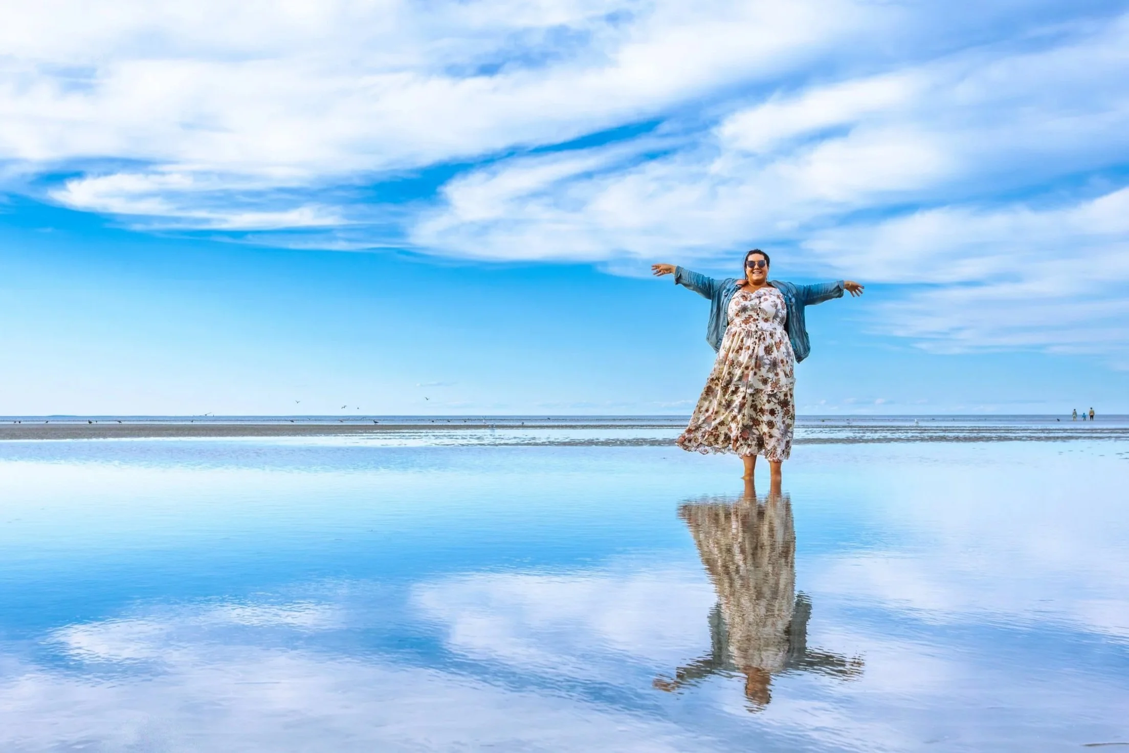 Woman in a floral dress with a denim jacket standing on a beach with water reflecting the sky and her, arms outstretched, under a partly cloudy blue sky.