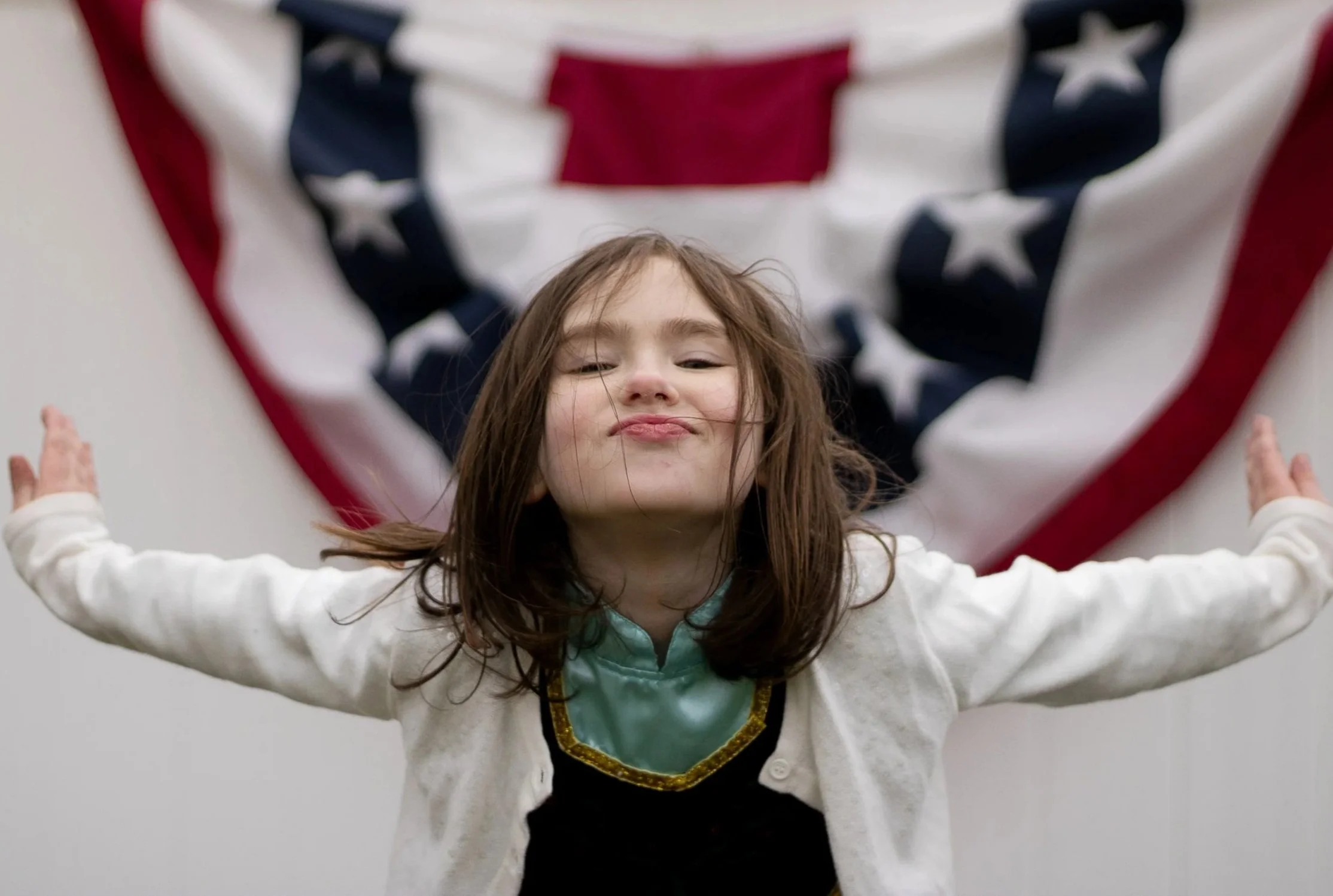 A young girl with brown hair making a confident face with arms outstretched in front of an American flag background.