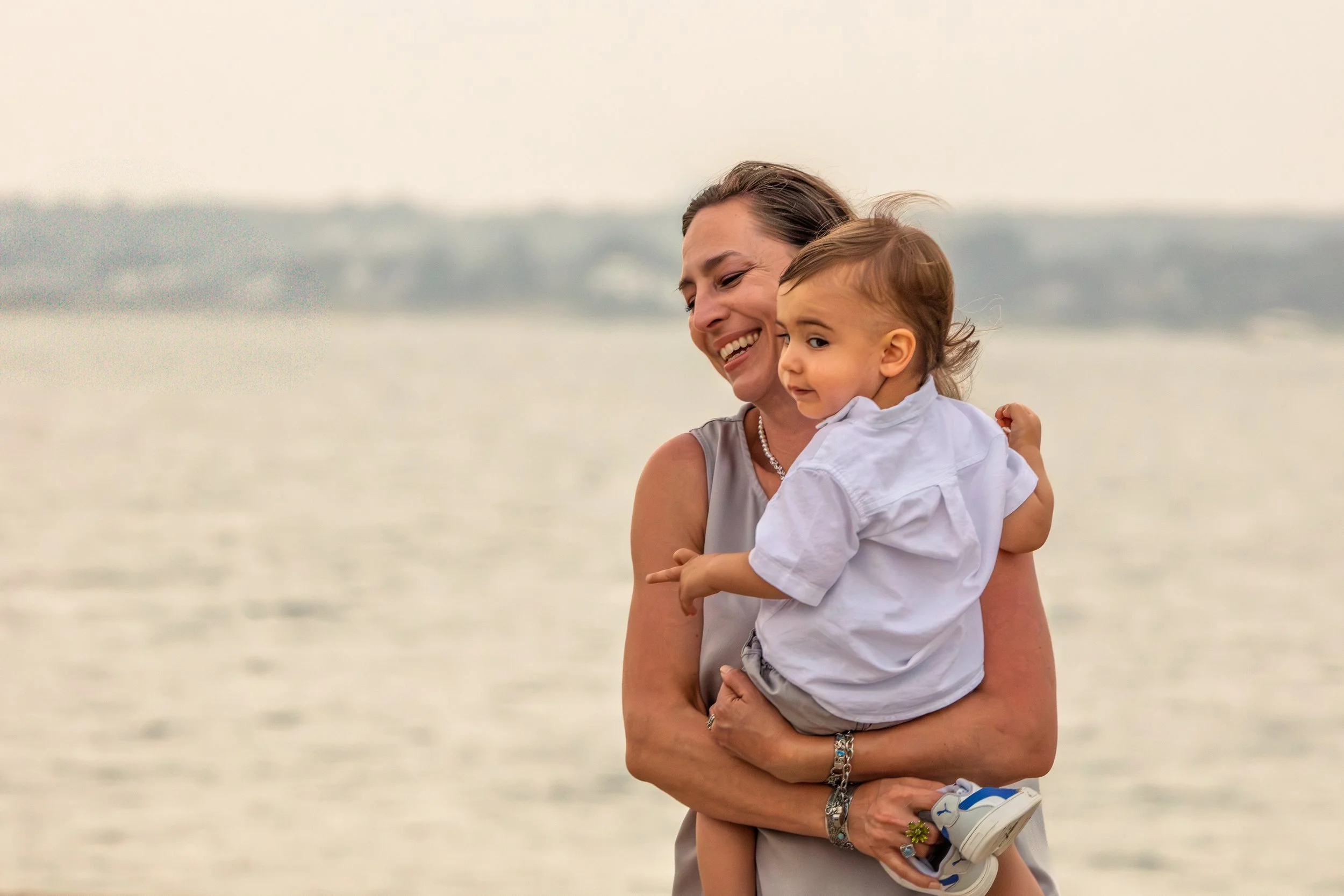 A woman holding a young child near a body of water with an overcast sky in the background.