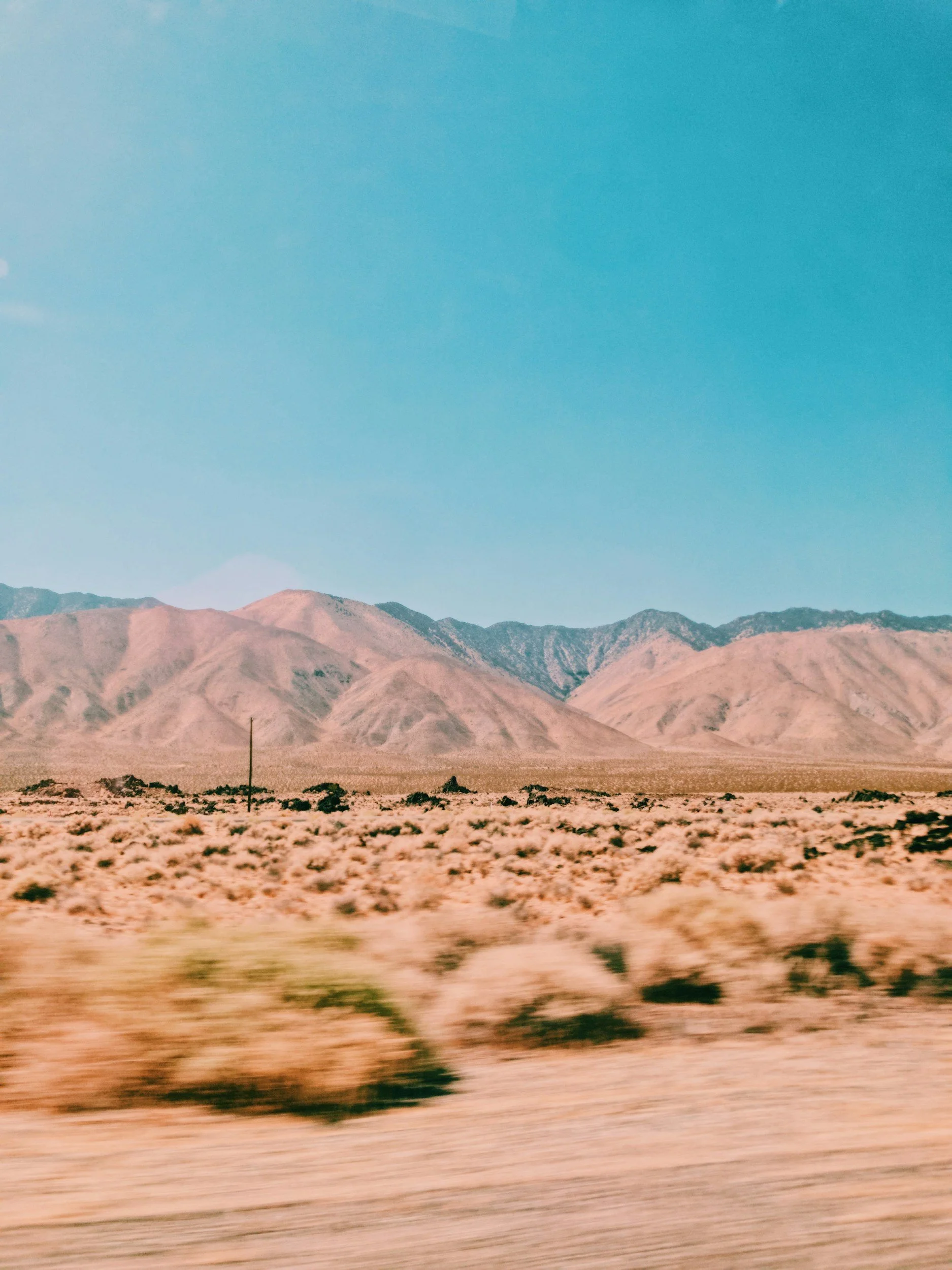 A desert landscape with dry, sandy terrain, scattered shrubs, distant mountains, and a clear blue sky.