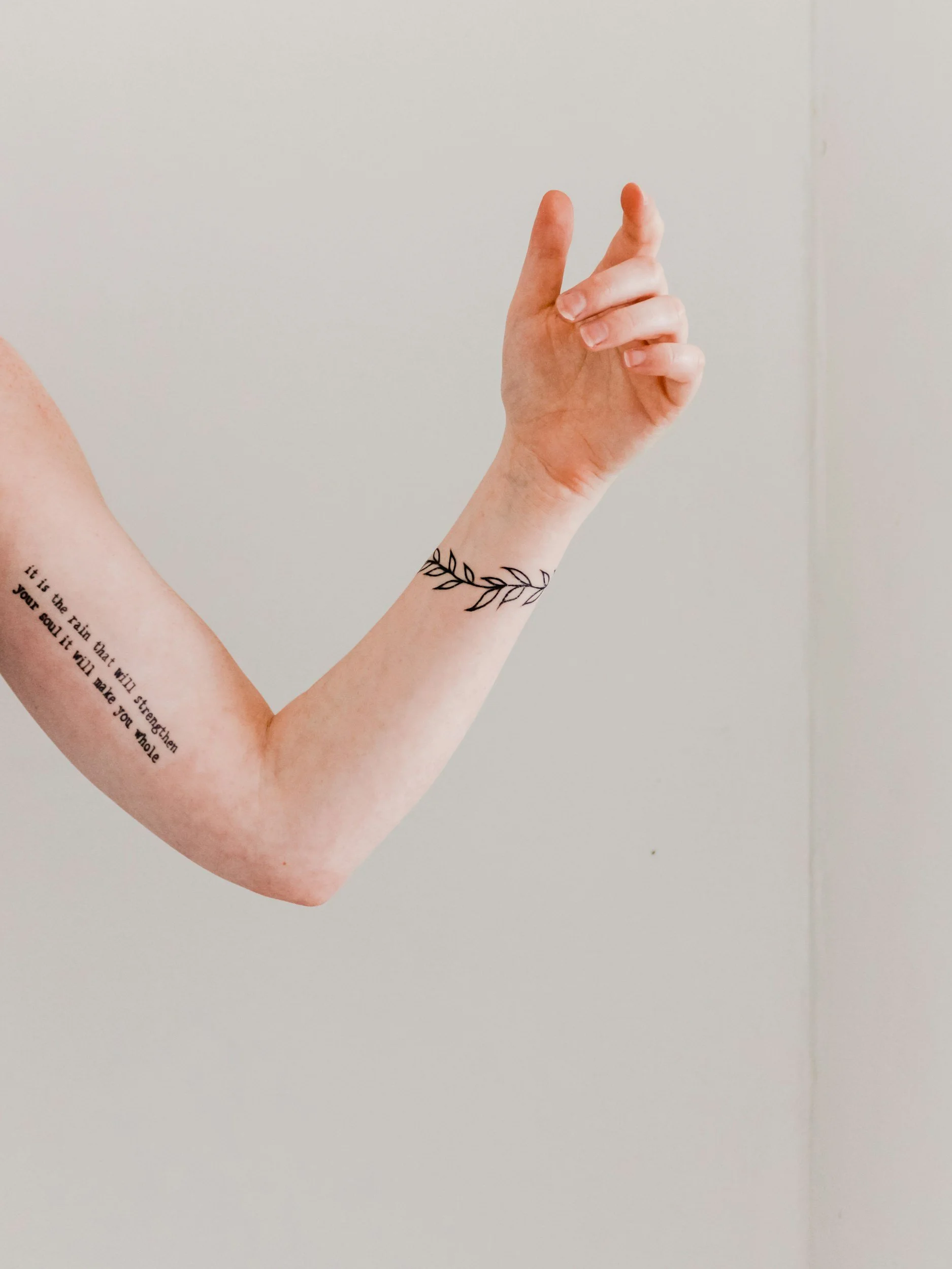 Close-up of a person's arm with a tattoo of a quote on the inner forearm and a black line drawing of a leaf on the wrist, against a plain background.