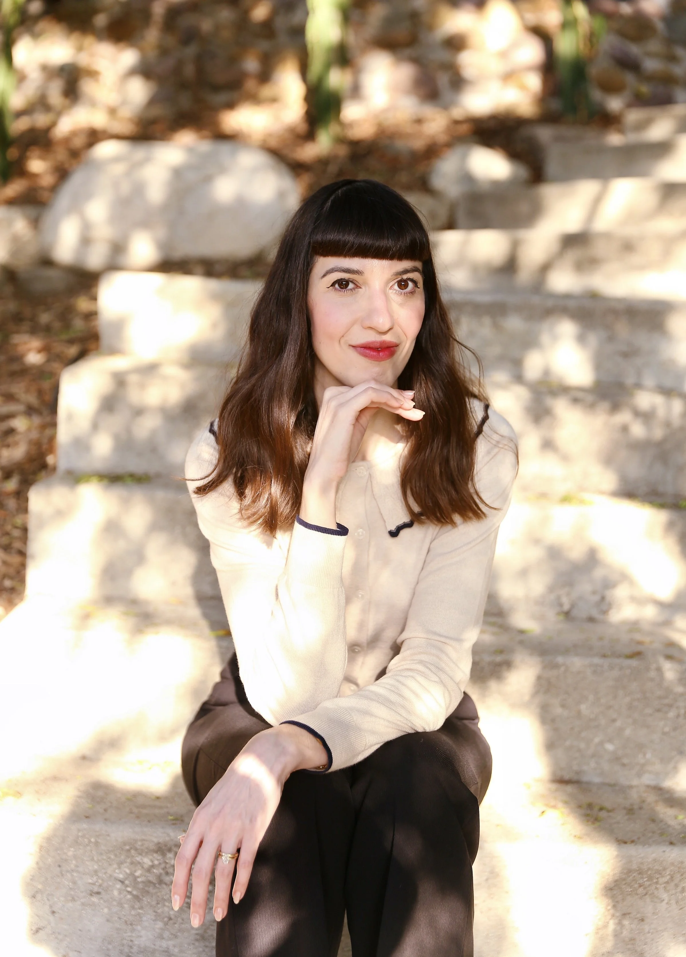 A woman with dark hair and bangs sitting on outdoor steps, wearing a cream-colored sweater and black pants, with her right hand resting on her chin and her left hand on her knee.