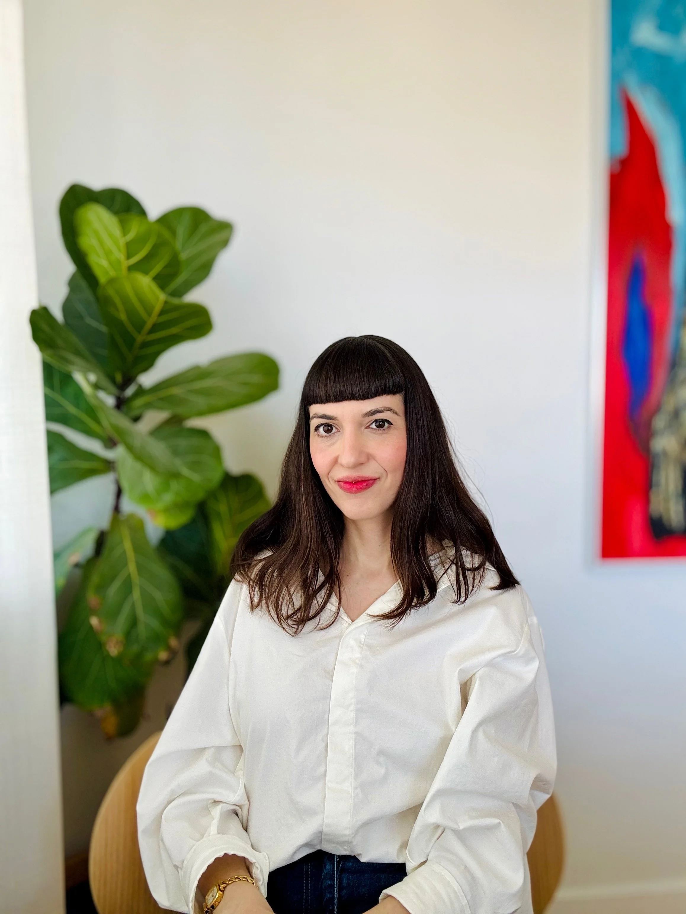 A woman with long brown hair and blunt bangs, wearing a white blouse, sitting in front of a large green plant with broad leaves and a colorful abstract painting on the wall.