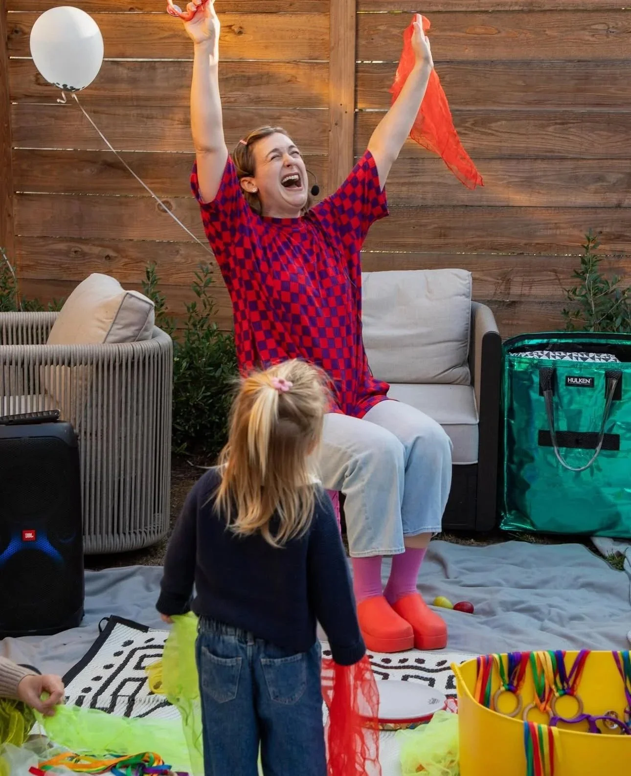 A woman with a short haircut and checkered shirt lifts her hands up as she sings. A small girl stands in front of her, looking up to her.