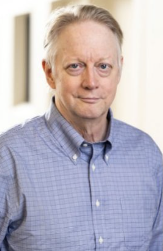Photo of David Dunning, a man with gray hair and a serious expression, wearing a light blue plaid button-up shirt, standing indoors with a blurred background.