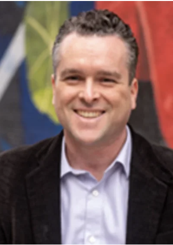 Photo of Jeff Schlegelmilch, a smiling man with short, curly gray hair wearing a light blue shirt and dark velvet blazer, standing outdoors.