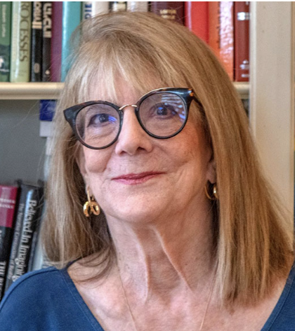 Photo of Elizabeth Loftus, a woman with shoulder-length red hair, glasses, and gold earrings, smiling in front of a bookshelf filled with books.