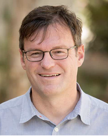 Photo of Craig Callender, a smiling man with brown hair wearing glasses and a light blue collared shirt outdoors with a blurred natural background.