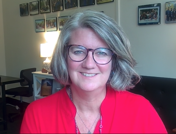 Photo of Katie Dyer, a woman with gray hair and glasses, smiling, wearing a red top, sitting in an office with framed photos on the wall behind her.