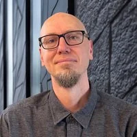 Photo of Rodney Schmaltz, a man with glasses and a goatee, wearing a gray shirt, standing outdoors in front of a gray stone wall and metal railing.