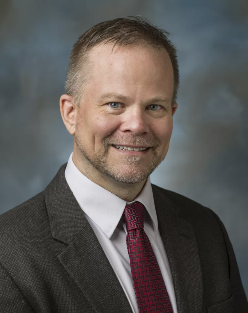 Photo of Kevin Folta, a man with light skin, short brown hair, blue eyes, and a beard, wearing a black suit, white shirt, and a red patterned tie, smiling at the camera.
