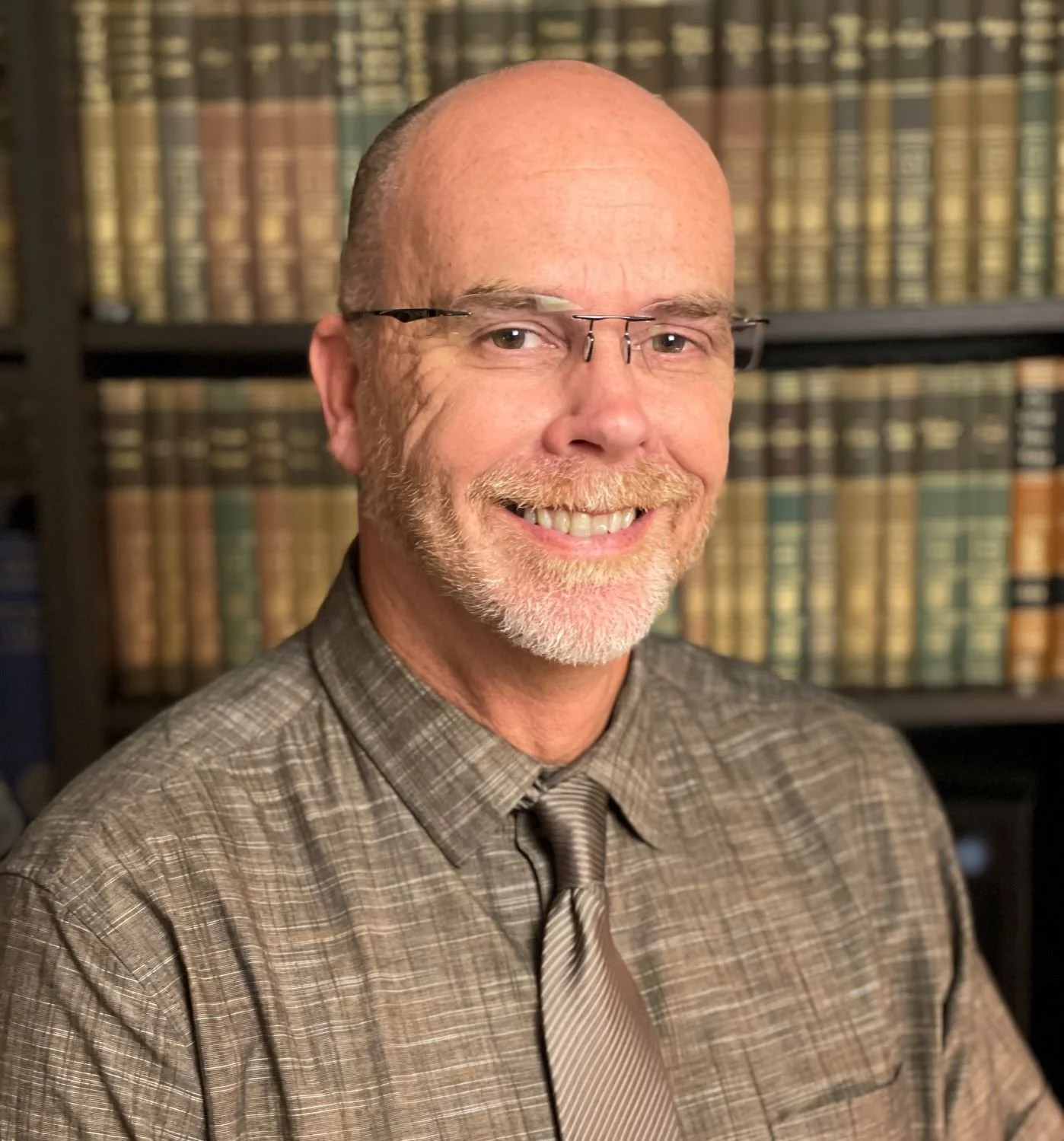 Photo of Ray Hall, a smiling man with glasses, a bald head, and a beard, wearing a plaid shirt and tie, in front of a bookshelf filled with books.