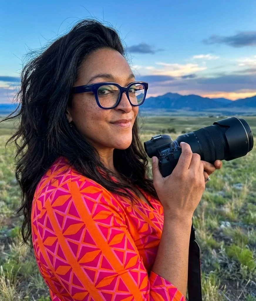 A woman with dark hair, glasses, and freckles smiling while holding a camera outdoors during sunset, with mountains and a vast landscape in the background.