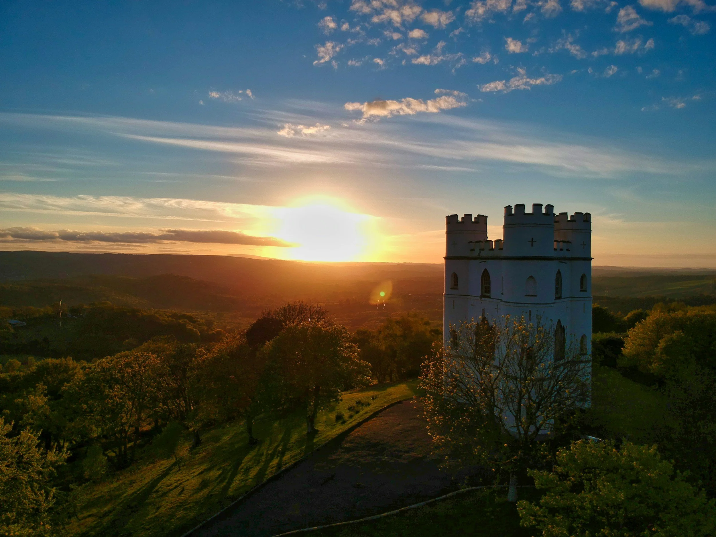 A scenic view of a white castle-like tower on a hill during sunset, with green trees and rolling hills in the background and a partly cloudy sky.
