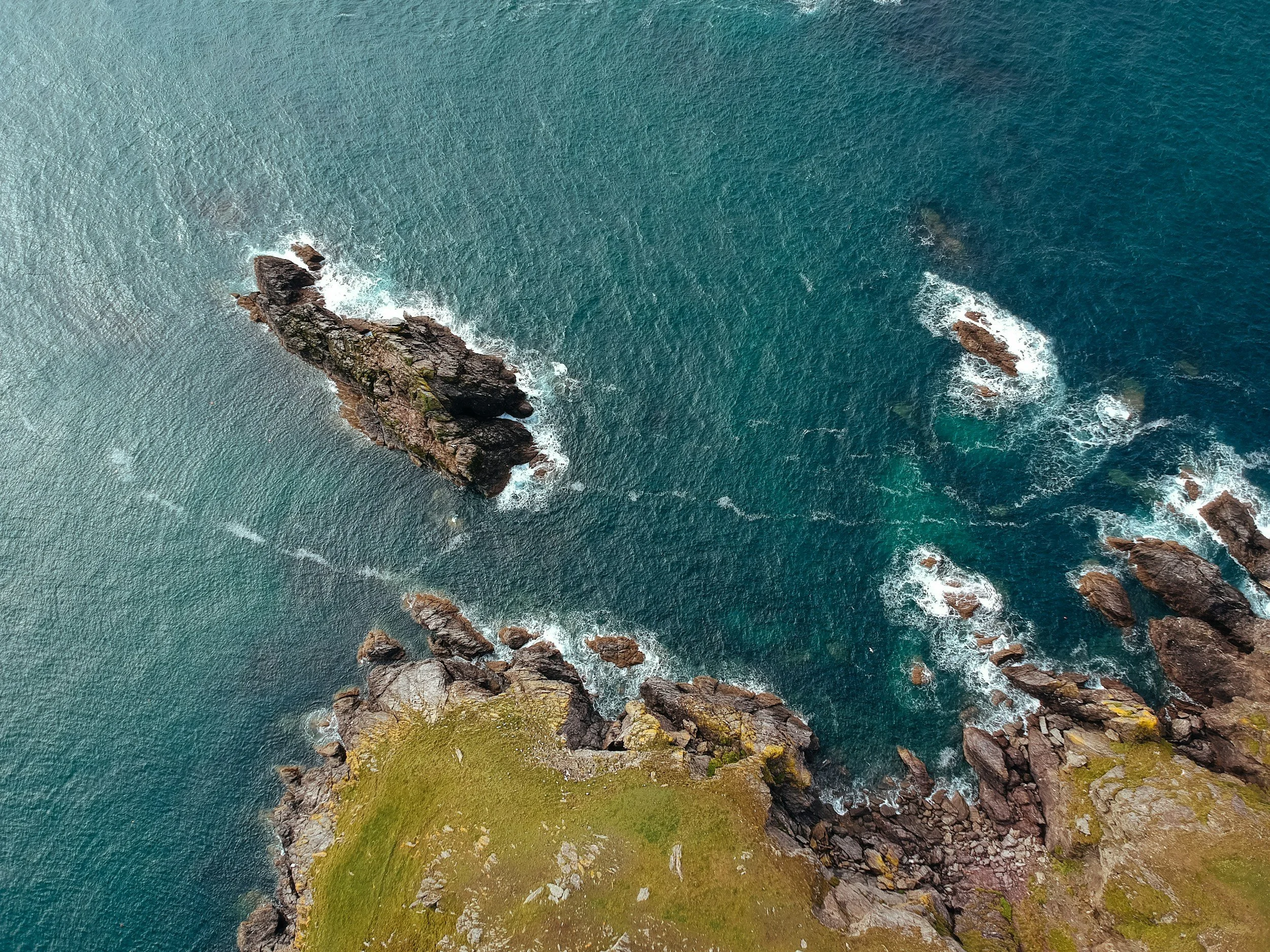Aerial view of rocky coastline with ocean waves crashing against rocks and a grassy area at the bottom.