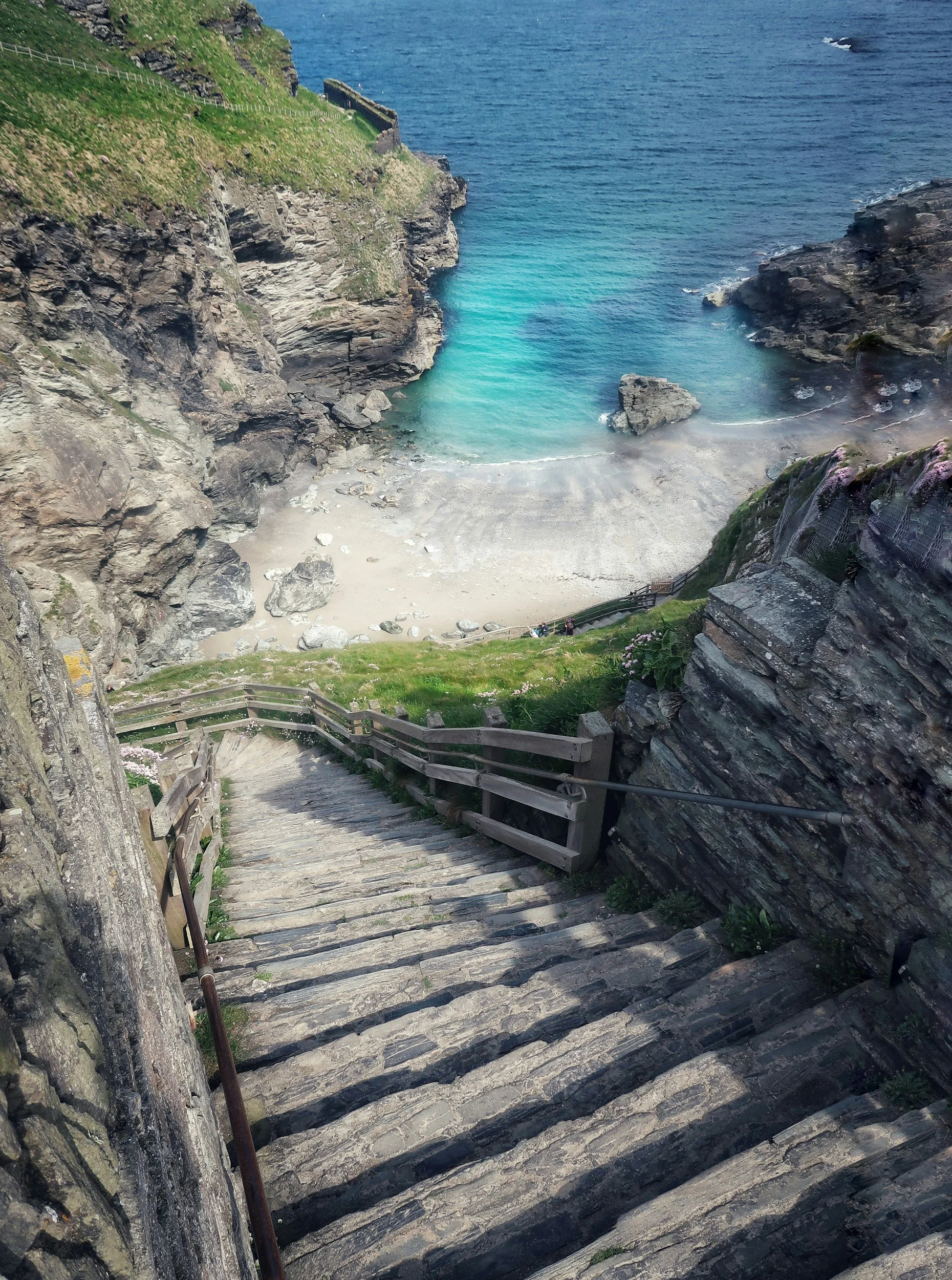 Stone stairway leading down a hillside towards a small beach with rocks, surrounded by cliffs and the ocean.