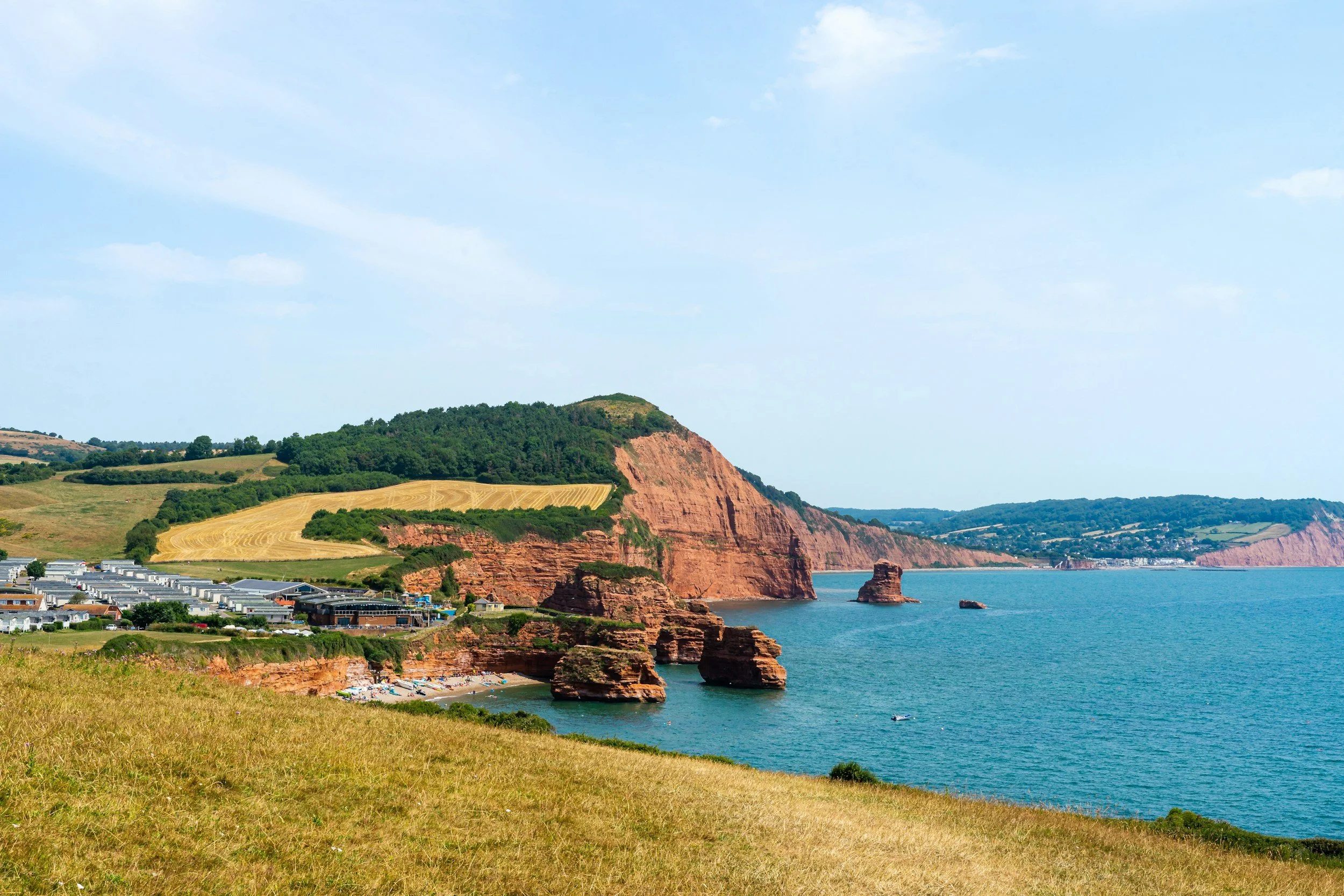 Coastal cliffs with red rock formations, a beach with umbrellas, and a green hillside under a partly cloudy sky.