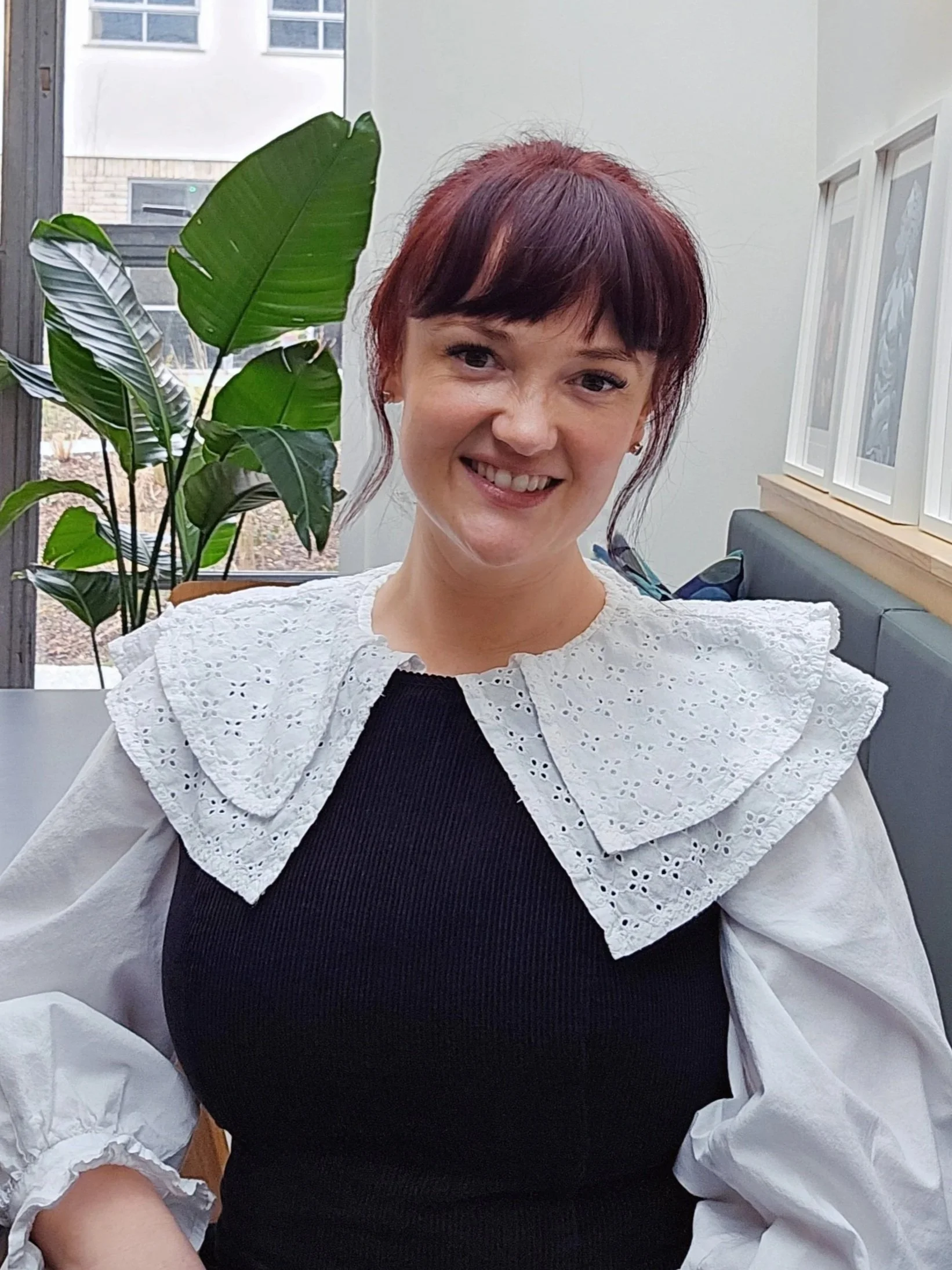 Young woman with short red hair smiling, wearing a white eyelet lace collar top with puffy sleeves, sitting indoors near a large plant with green leaves.