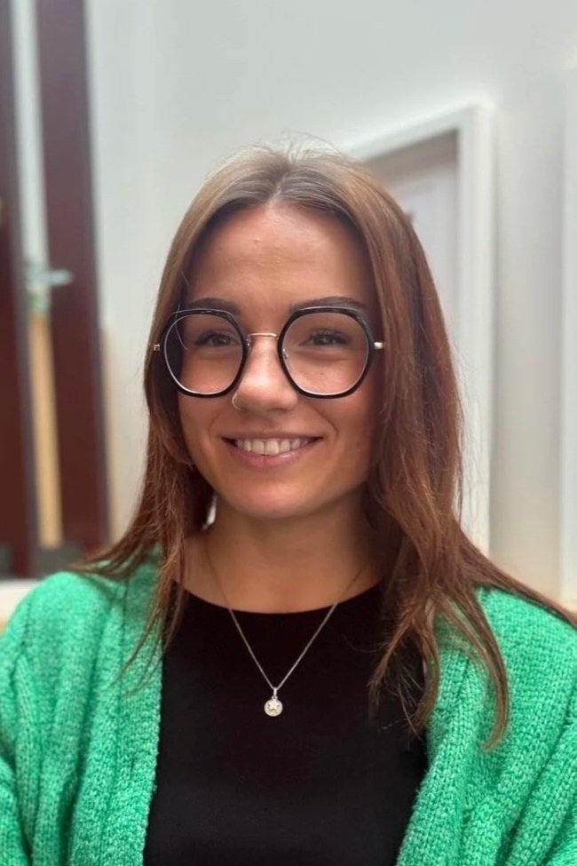 A woman with shoulder-length brown hair wearing large black-rimmed glasses, a black top, a green cardigan, and a silver necklace, smiling indoors.