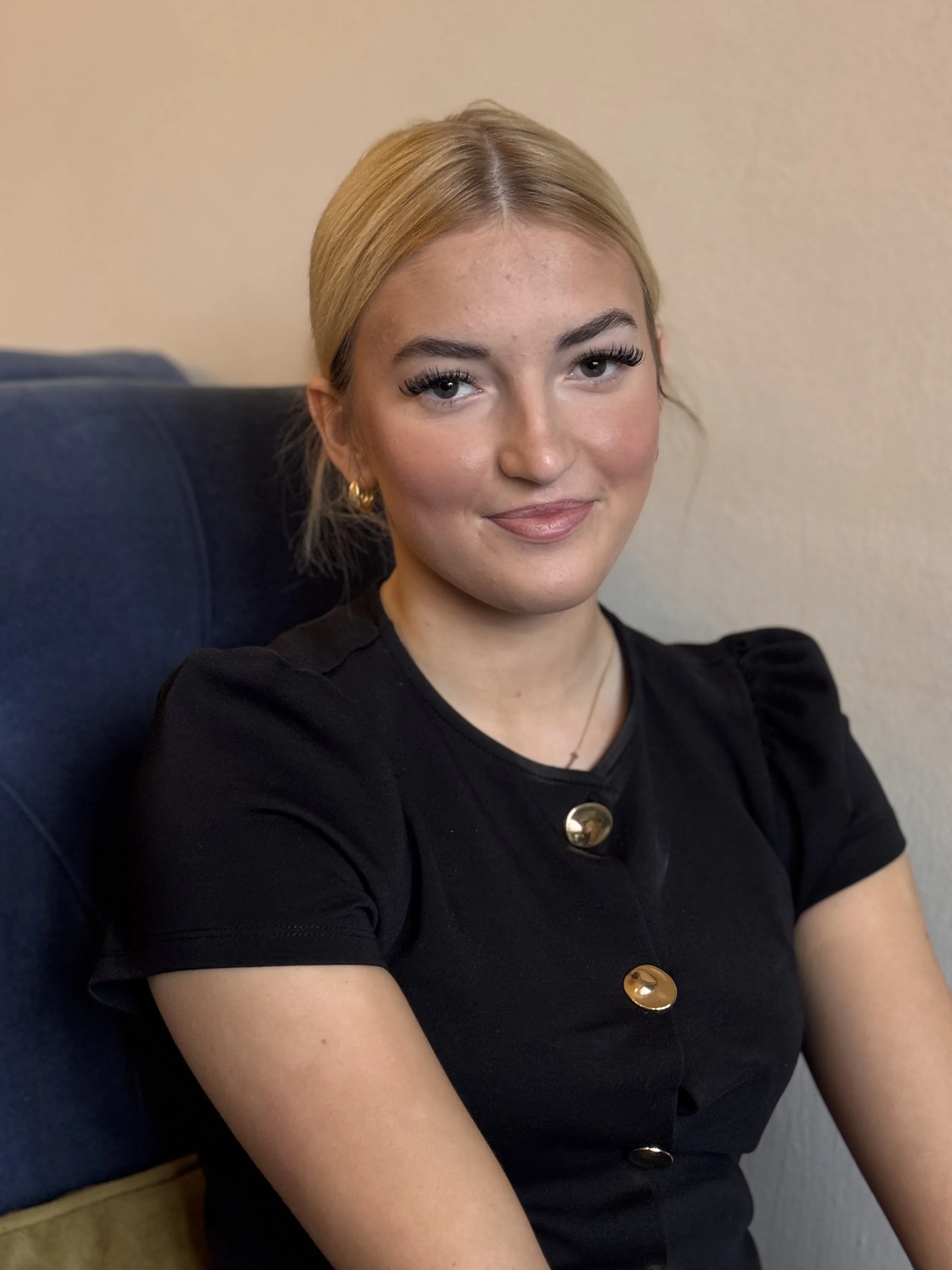 A young woman with blonde hair, wearing a black top with gold buttons, sitting on a dark blue chair, smiling softly at the camera.