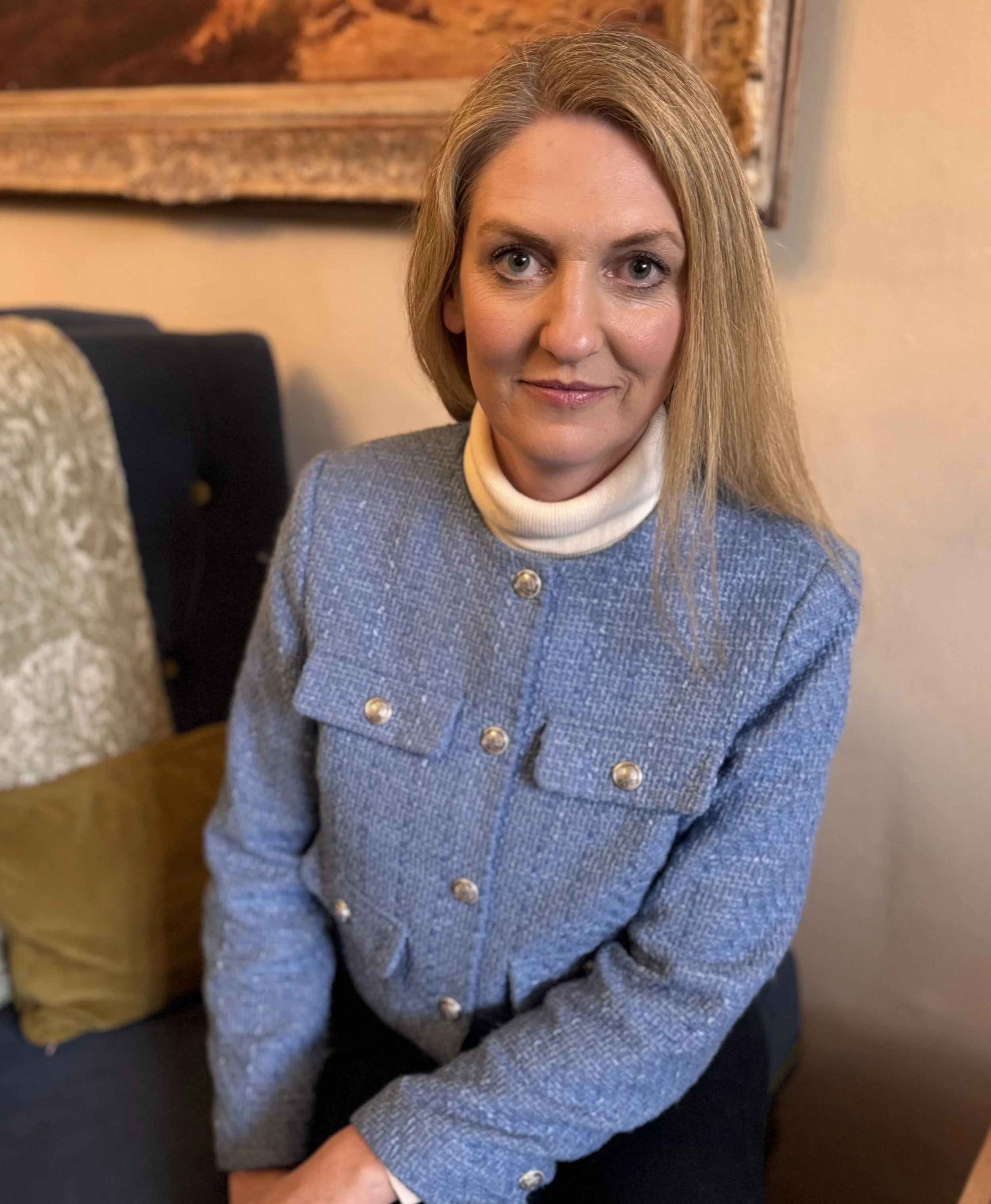 A woman with shoulder-length blonde hair, wearing a light blue tweed jacket with silver buttons and a white turtleneck, sitting on a dark-colored sofa in a warmly lit room.