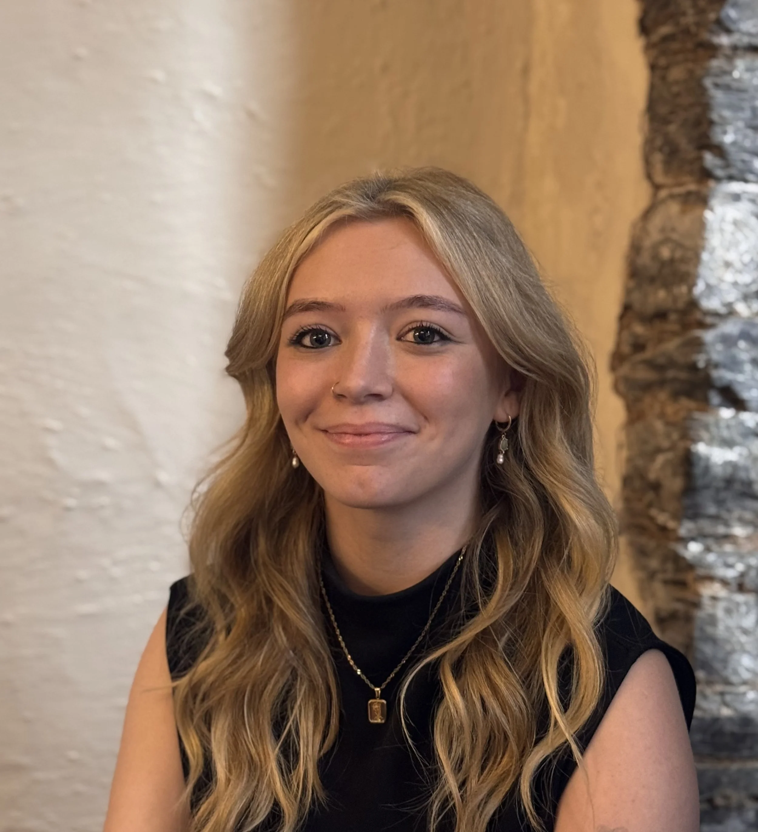 A young woman with long wavy blonde hair, wearing a black top, pearl earrings, and layered gold necklaces, smiling softly inside a room with a textured white wall on her left and a stone wall behind her.