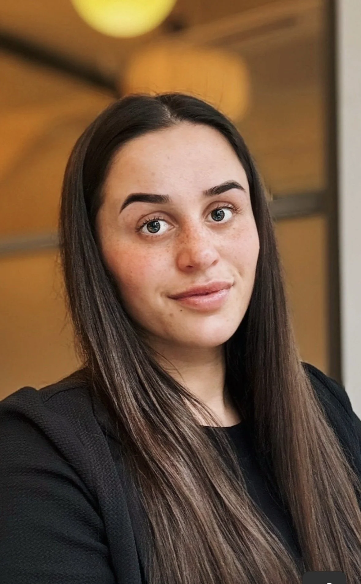 A young woman with long brown hair and freckles, wearing a black top, looking at the camera with a slight smile, in an indoor setting with a wooden background and soft lighting.