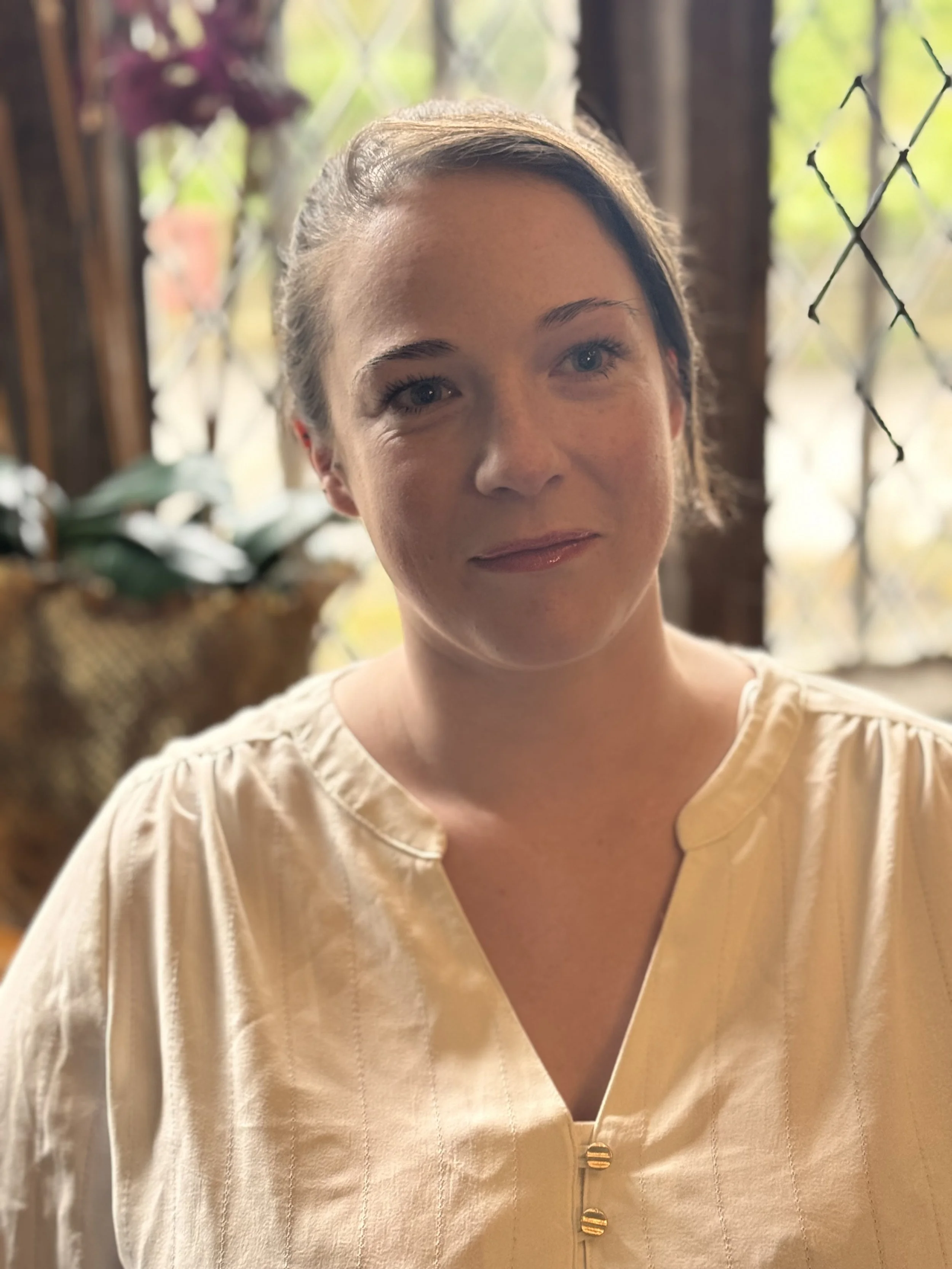A woman with short dark hair in a beige blouse, standing indoors with a blurred background of plants and daylight.