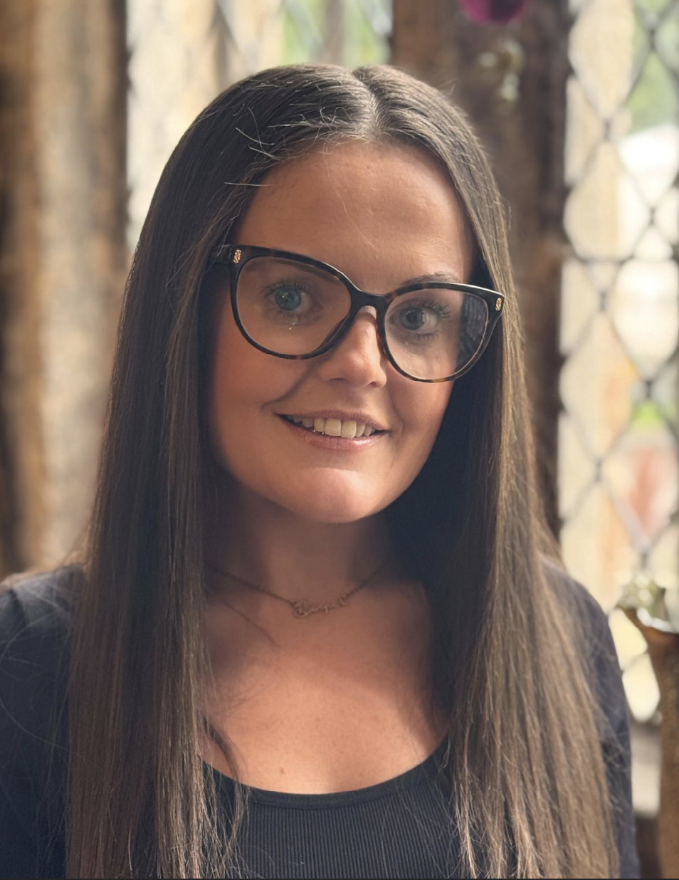 A woman with long brown hair wearing black-framed glasses and a black top, smiling at the camera, with a colorful background and wooden elements.