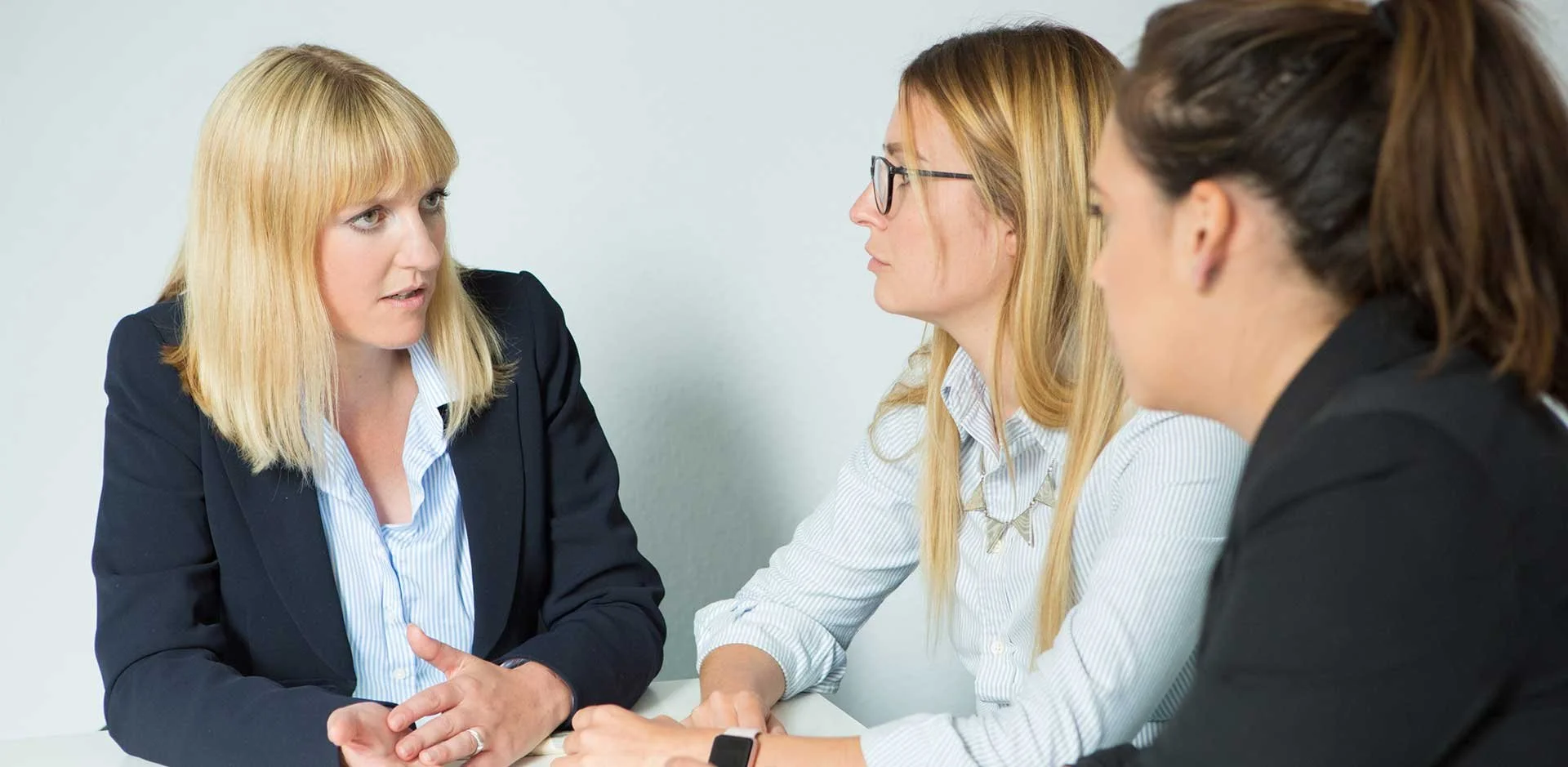 Three women engaged in a serious conversation at a meeting, sitting at a white table against a plain background.