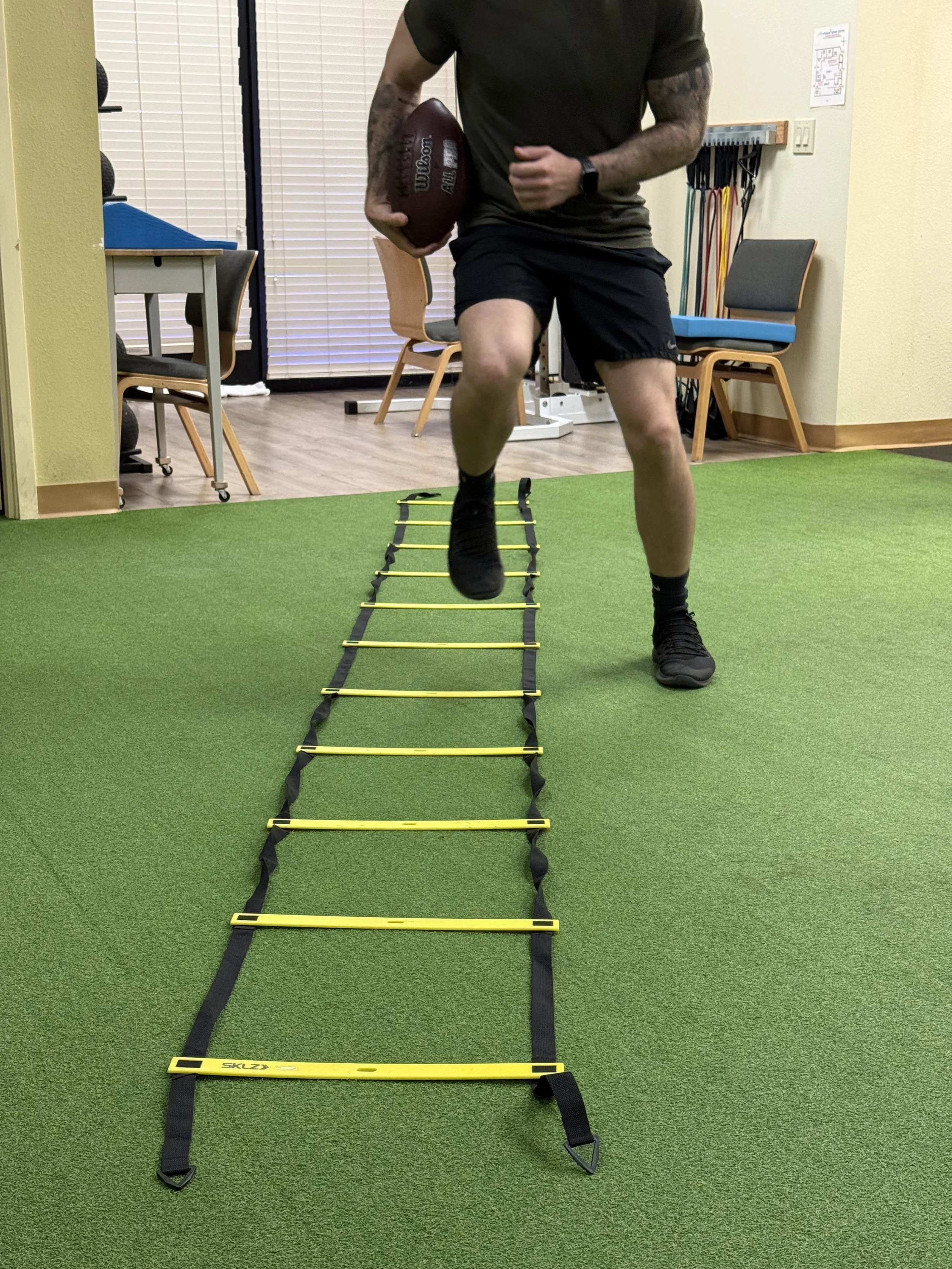 A fitness trainer or physical therapist crouched down and explaining something to a person standing on a yoga mat in a gym or therapy room. The person standing has tattoos on their legs and is barefoot.